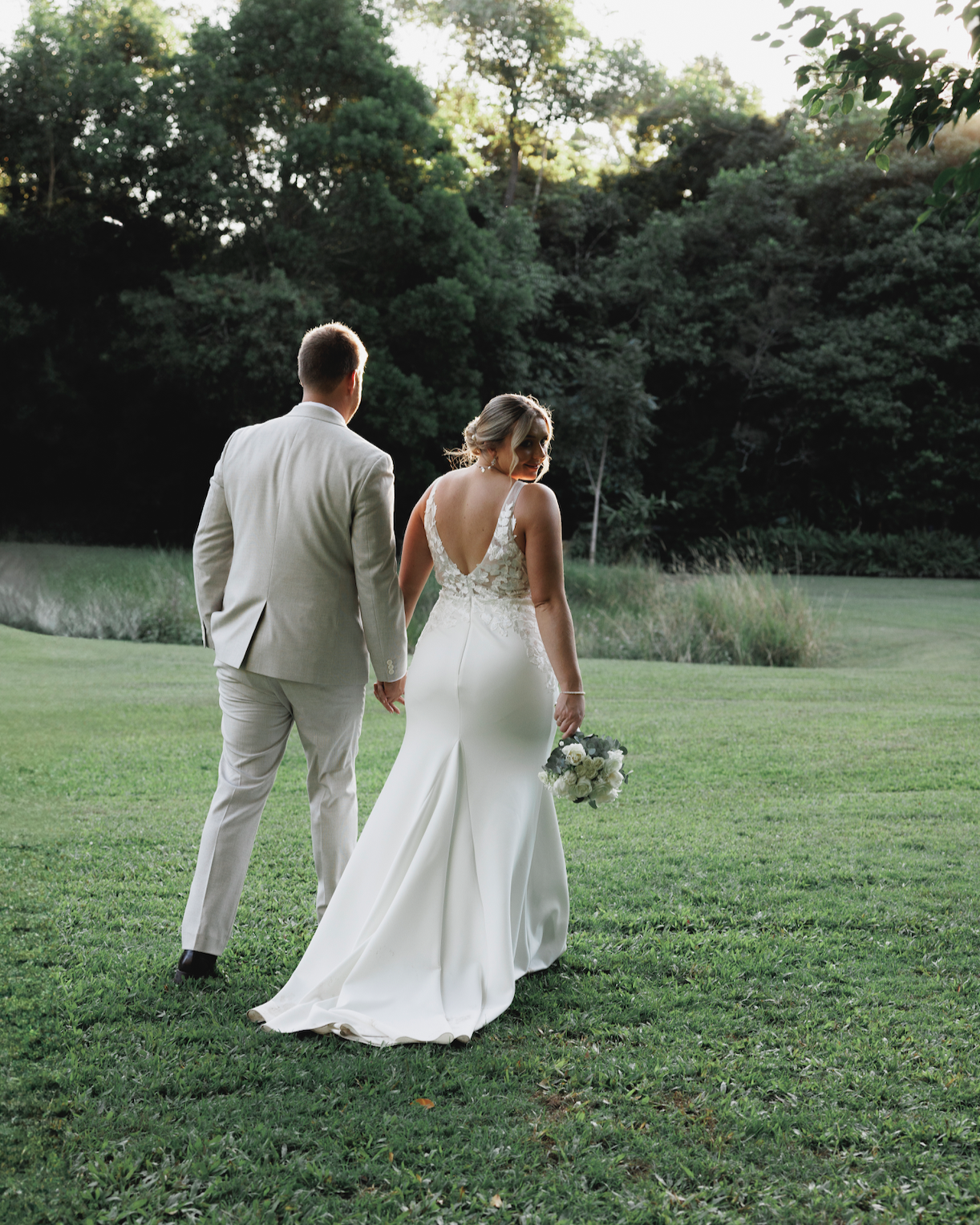 A bride and groom walking hand in hand on a grassy field, with trees in the background, during sunset or late afternoon. The bride is wearing a white wedding gown and holding a bouquet, while the groom is in a light-colored suit.