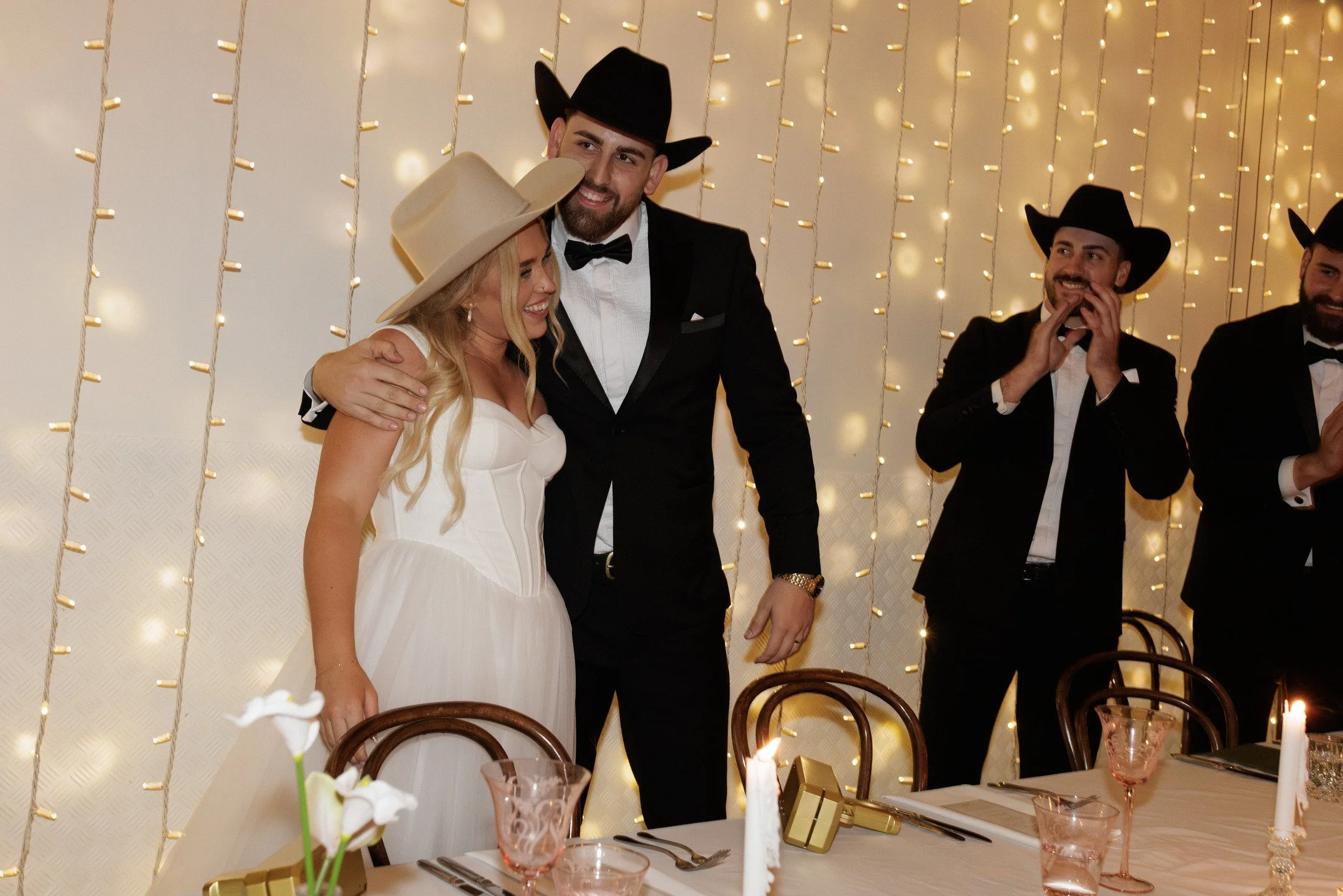 A wedding reception with a bride in a white wedding dress and a tall beige hat, and a groom in a tuxedo and black cowboy hat, smiling and embracing each other. Three men in tuxedos and cowboy hats are in the background near a decorated wall with stri