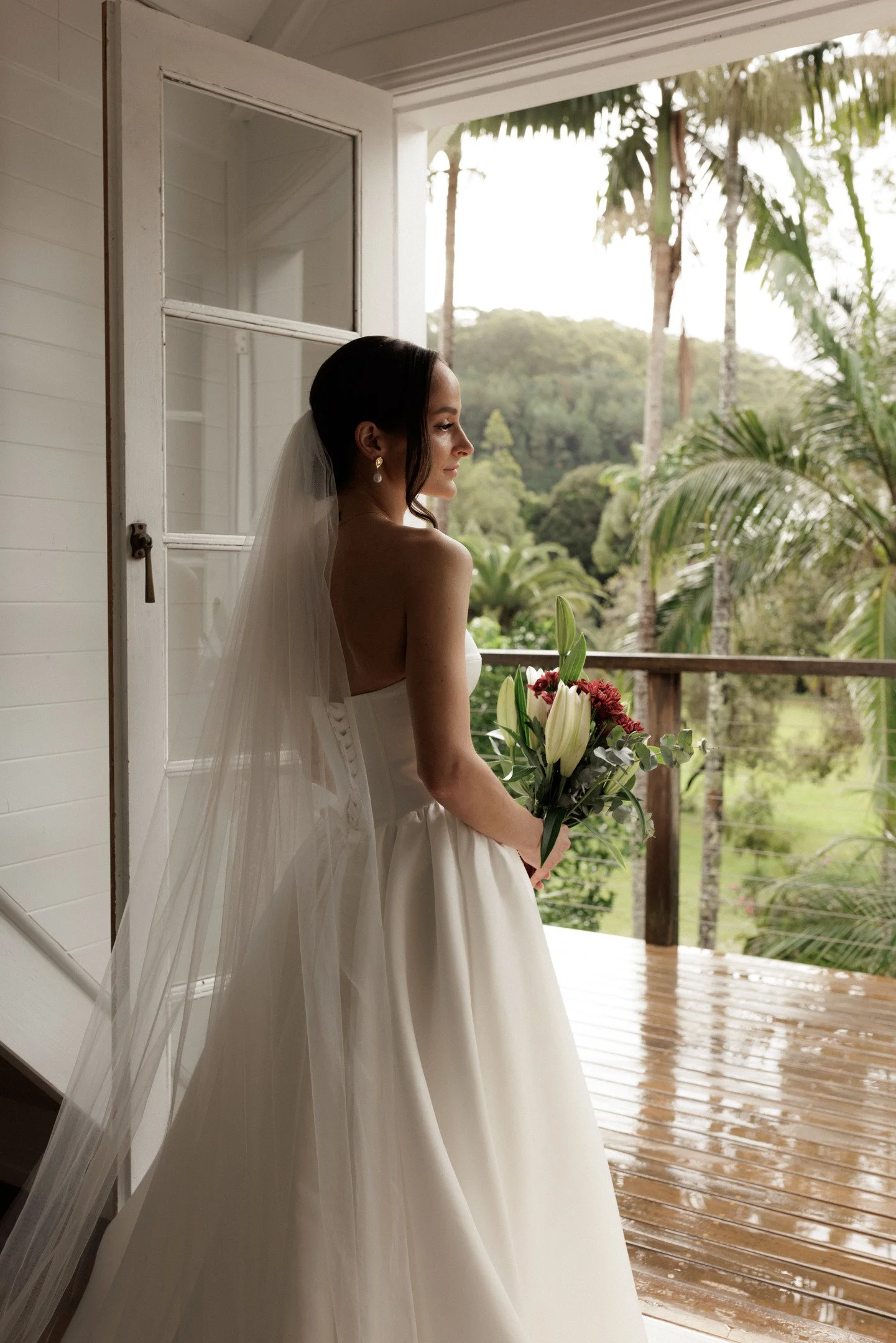 A bride standing on a porch, holding a bouquet of white lilies and pink flowers, with lush greenery and tall palm trees in the background.