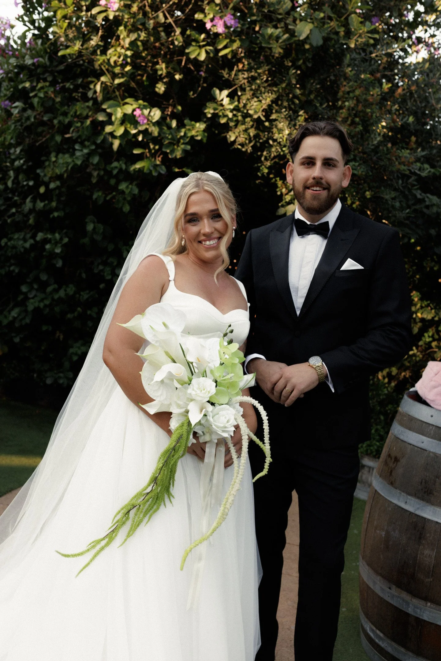 A bride and groom at their wedding outdoors, smiling, with a large green leafy bush with purple flowers in the background. The bride wears a white wedding gown with a veil and holds a bouquet of white flowers. The groom is dressed in a black tuxedo w
