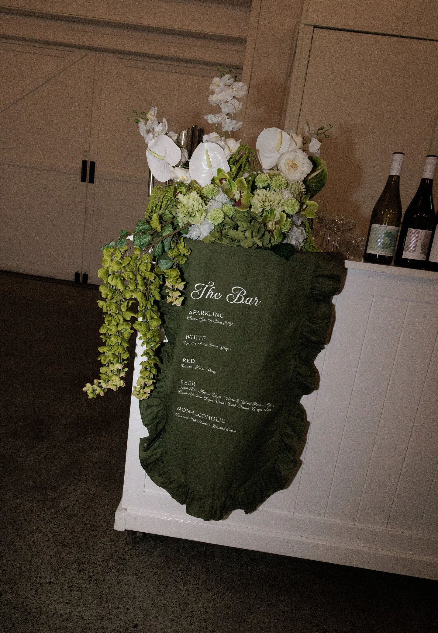 A floral arrangement on a bar counter with white orchids, green hydrangeas, and other greenery. The bar menu is visible hanging from the arrangement with options for sparkling, white, red, beer, and non-alcoholic beverages.