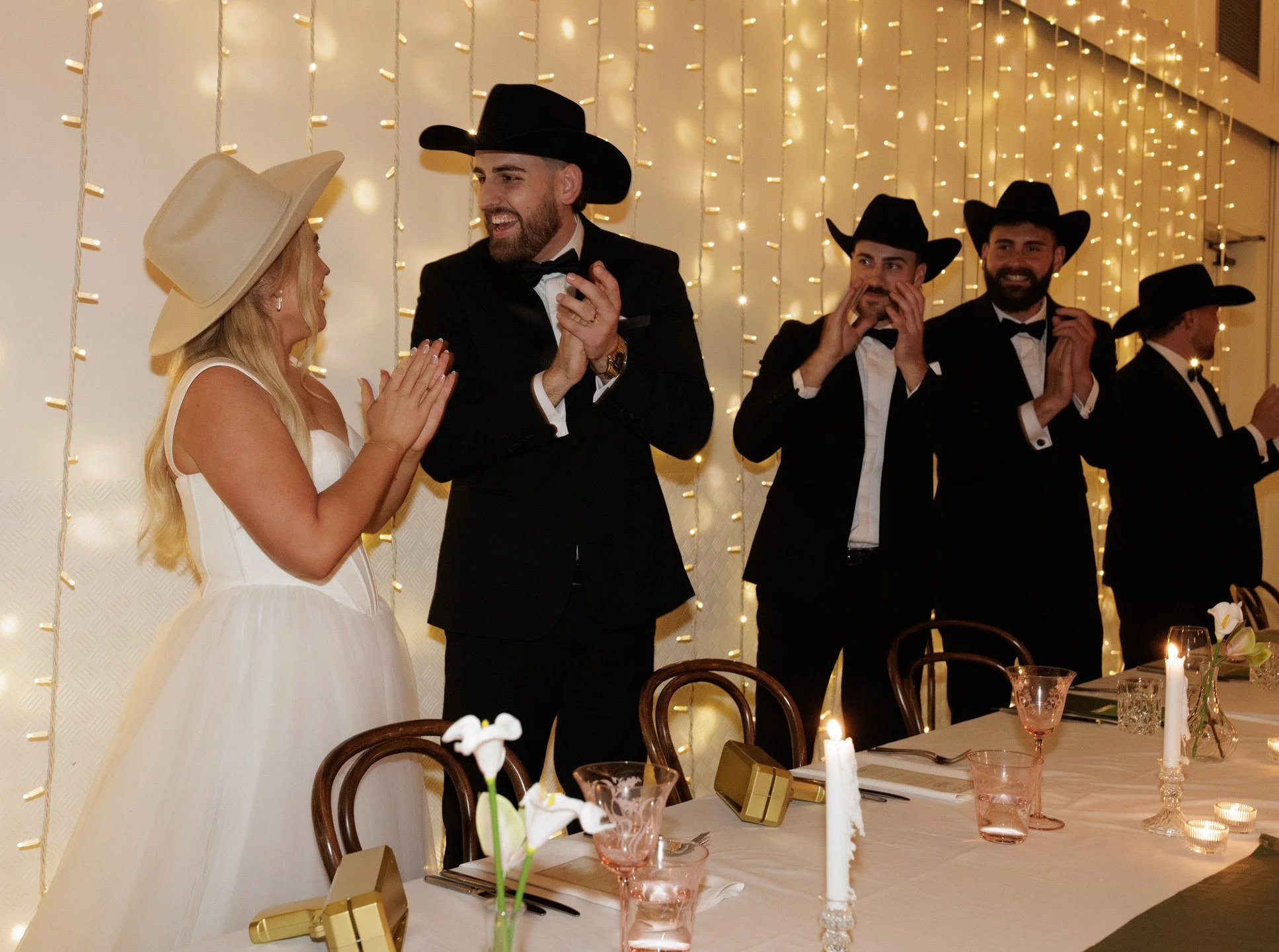 People enjoying a celebration at a formal event, dressed in tuxedos and a wedding dress, with string lights in the background. The woman is wearing a white dress and a large hat, while the men are in black tuxedos and cowboy hats.