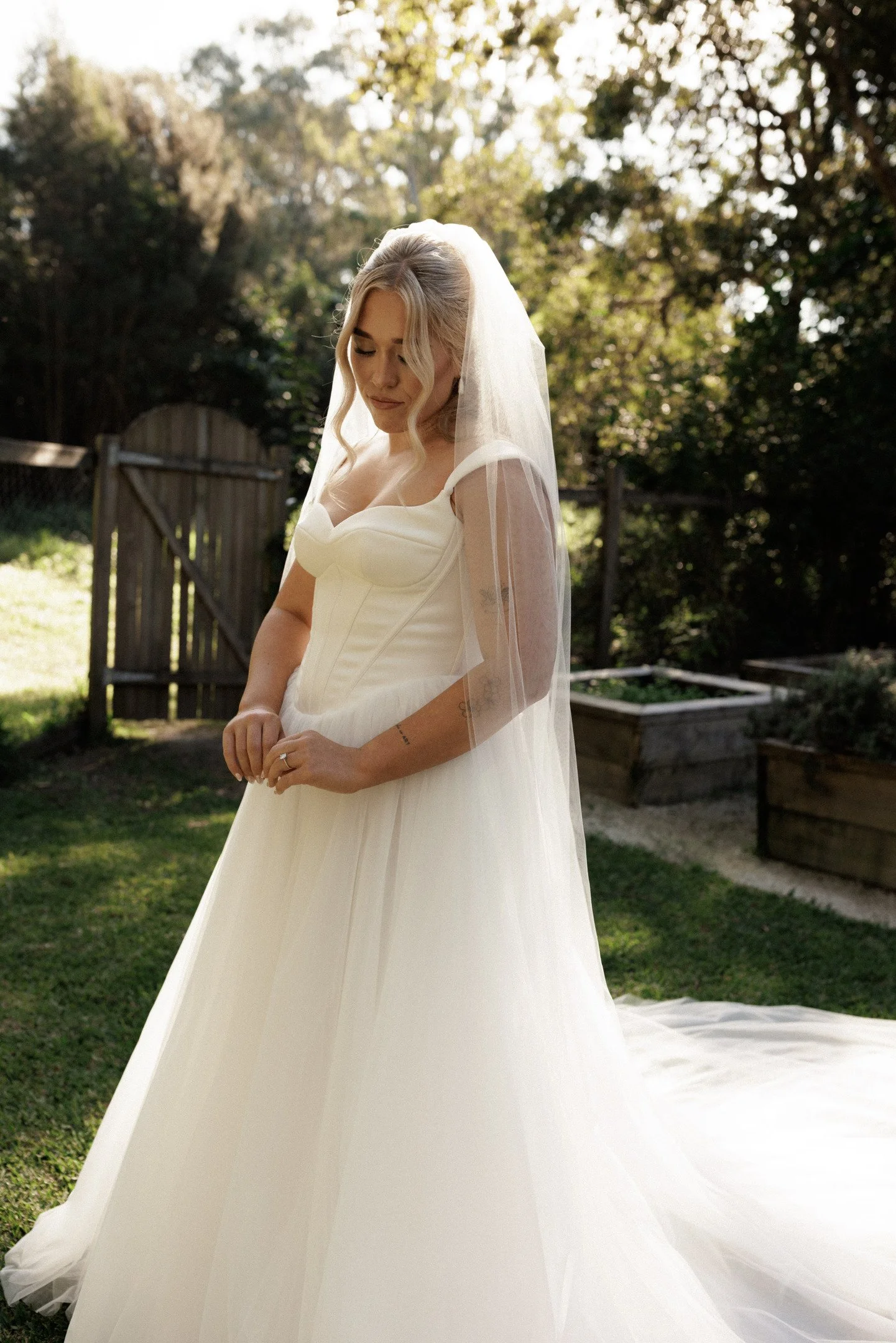 A bride wearing a white wedding dress and veil, standing outdoors with greenery and trees in the background.
