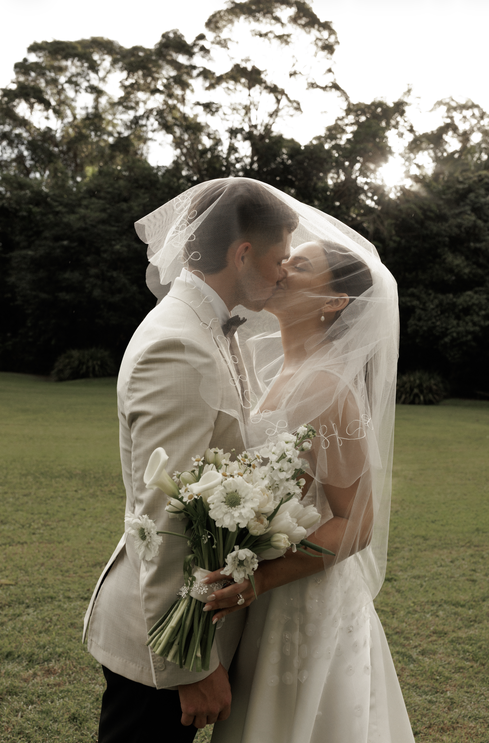 A bride and groom kissing outdoors, with the bride holding a bouquet of white flowers and wearing a wedding dress and veil, and the groom in a light-colored tuxedo, during sunset with trees in the background.