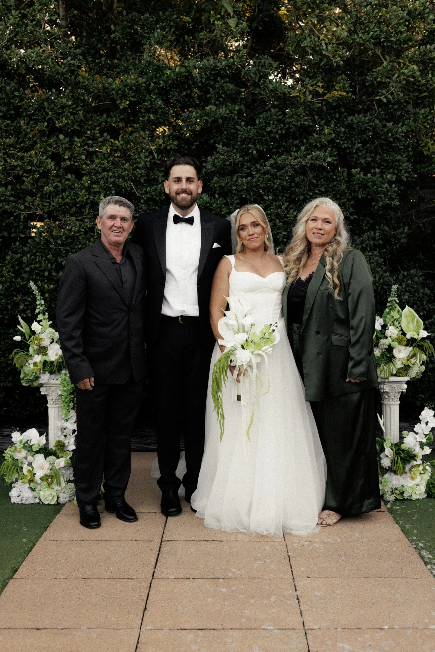 A group of four people at a wedding, standing on a paved area with floral decorations on either side, in front of a green leafy background. The man on the left is wearing a black suit, the man next to him is wearing a tuxedo with a bow tie, the bride