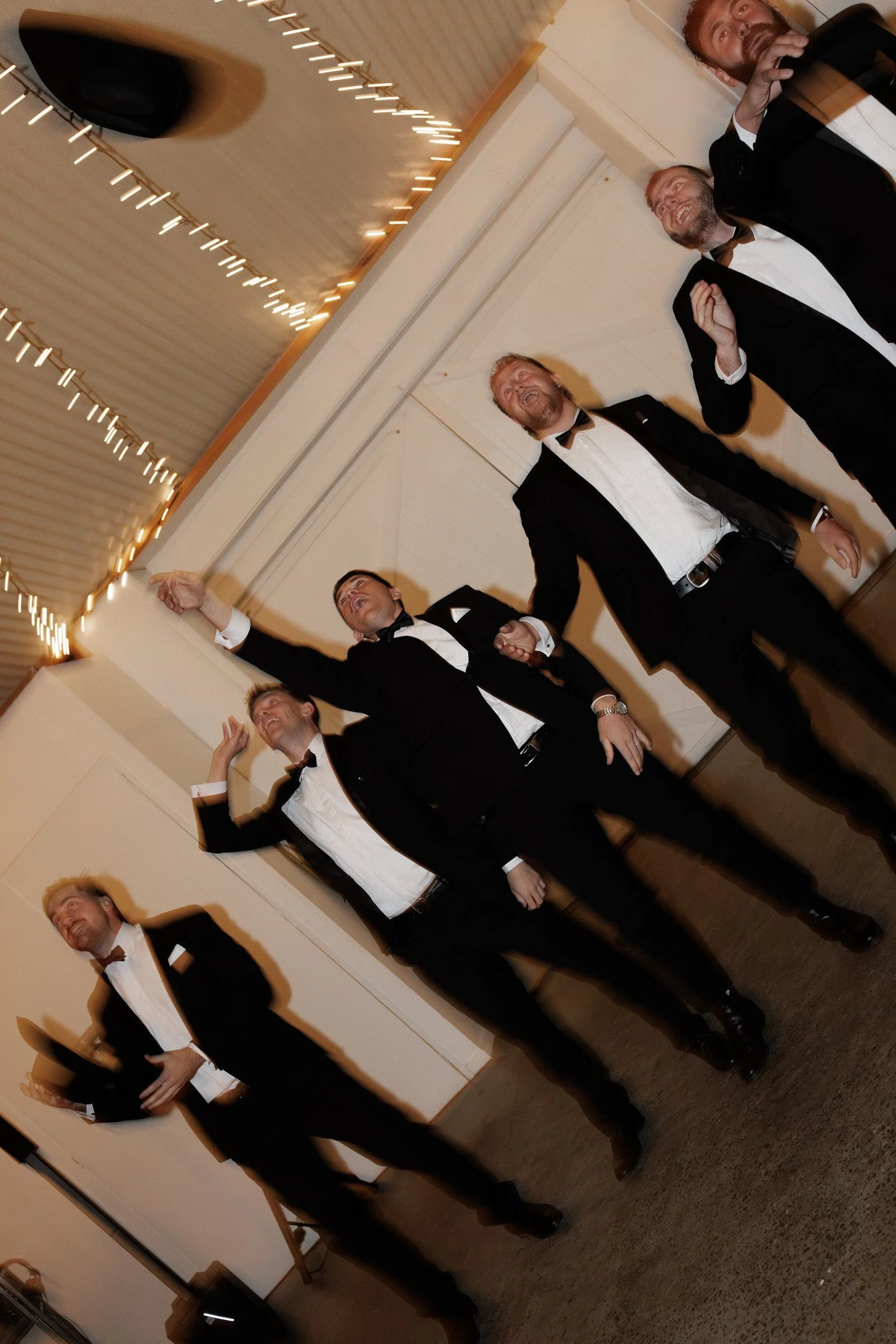A group of five men dressed in formal tuxedos with bow ties, standing together indoors, laughing and smiling at a social event, with a ceiling decorated with string lights.