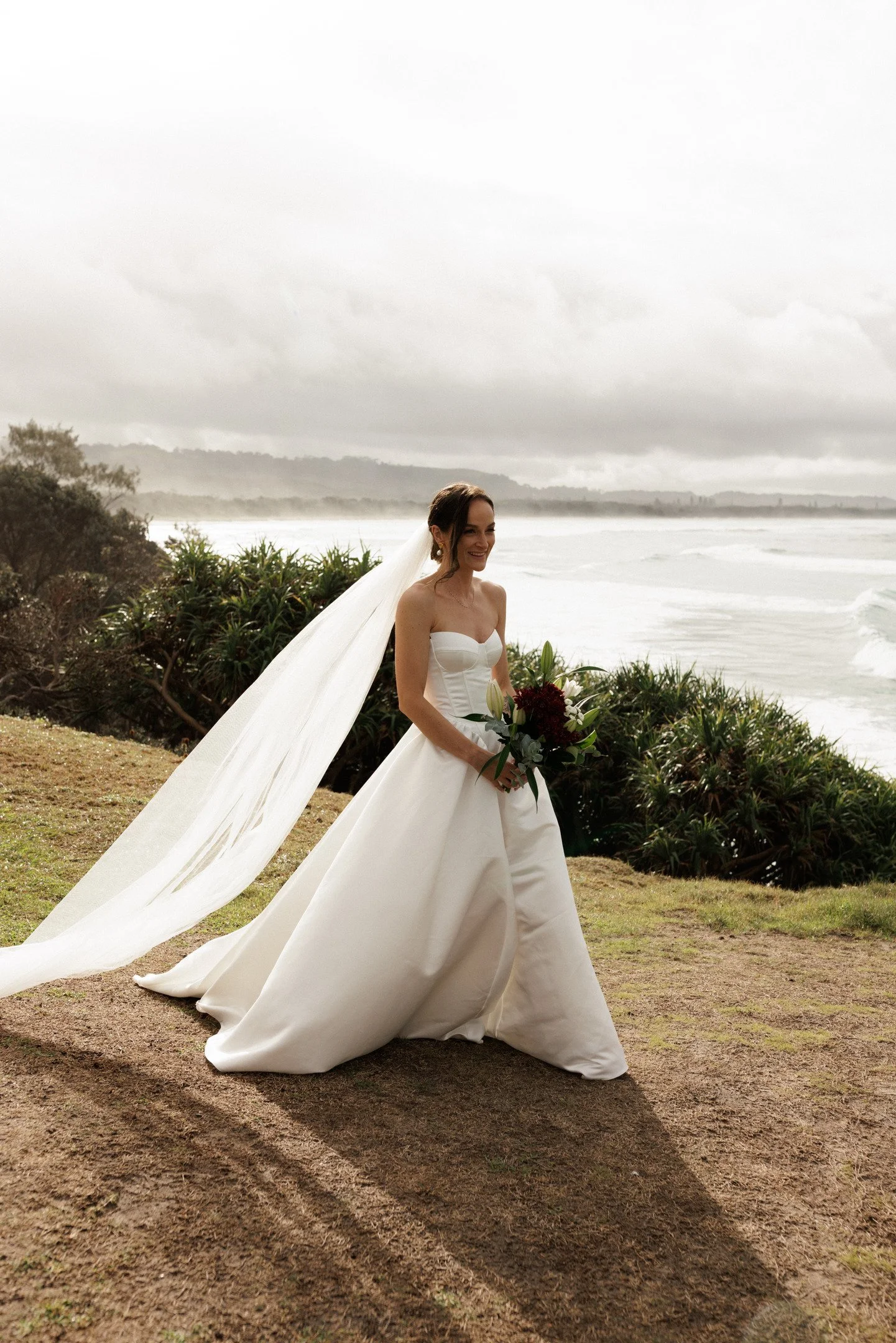 A woman in a white wedding gown standing outdoors near the coast with greenery and a cloudy sky in the background, holding a bouquet of red and white flowers.