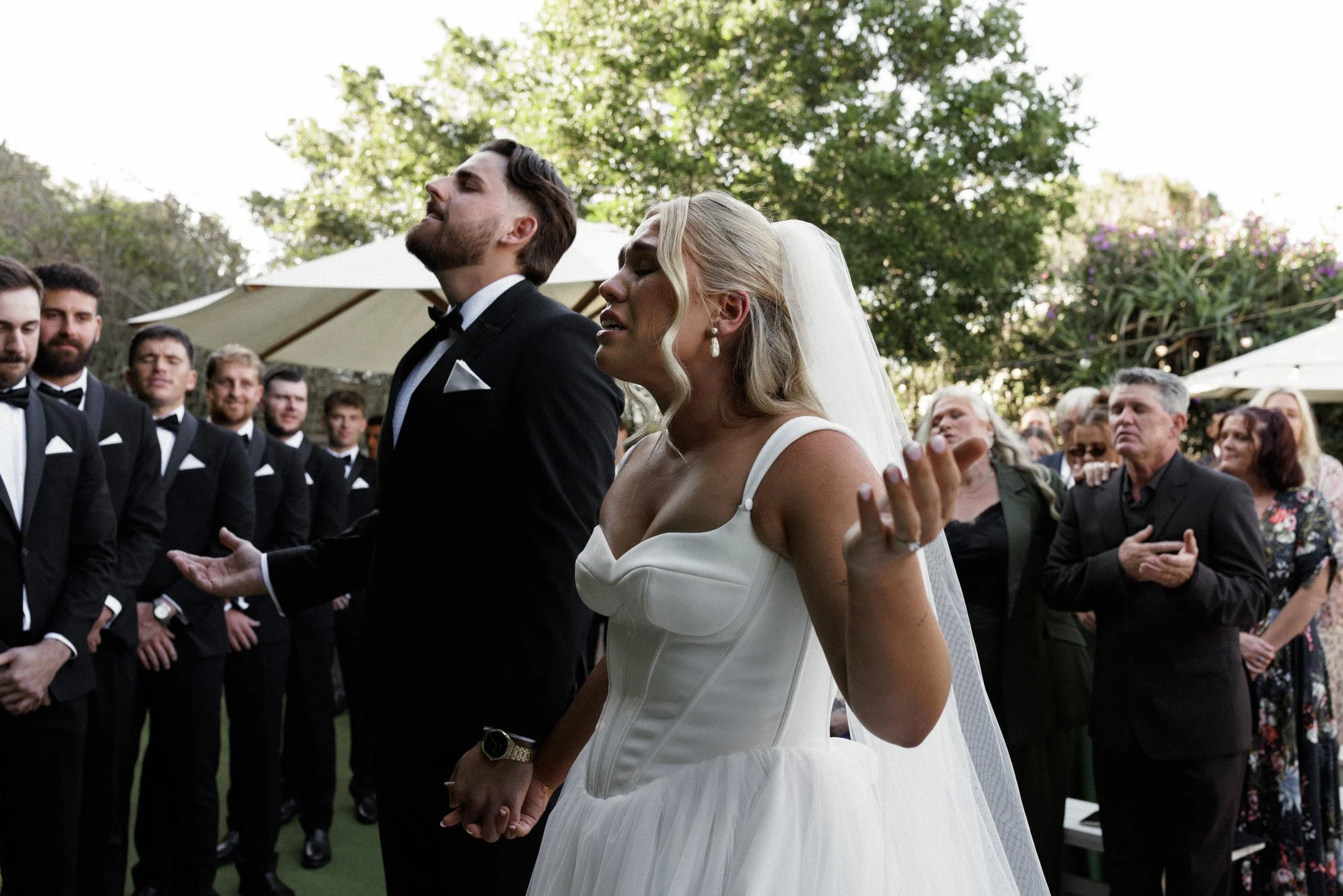 A bride and groom hold hands and kneel in prayer at their outdoor wedding ceremony surrounded by guests dressed in formal attire.