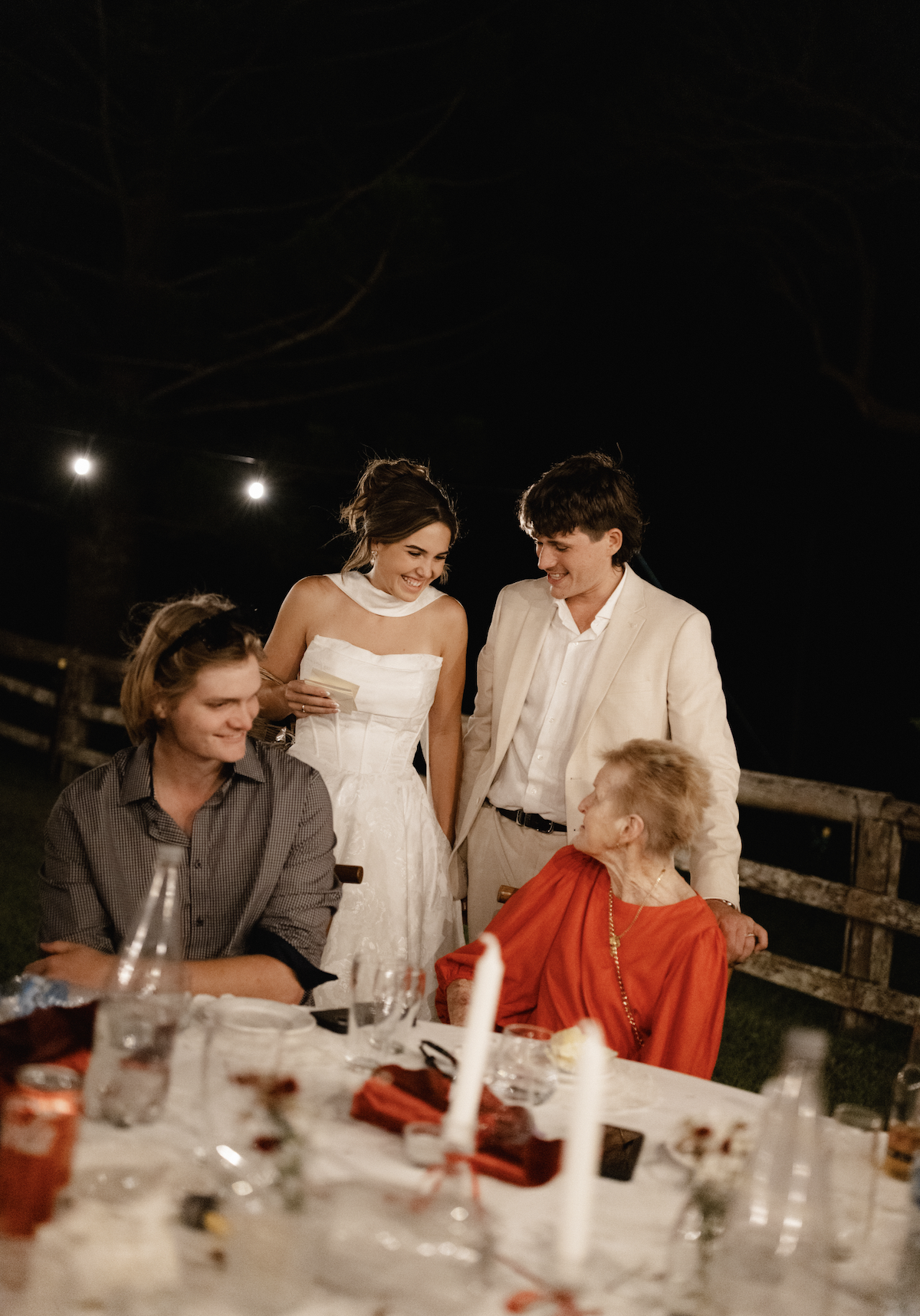 People celebrating at a dinner table outdoors at night, including a woman in a white dress, a man in a cream suit, and an elderly woman in a red dress.