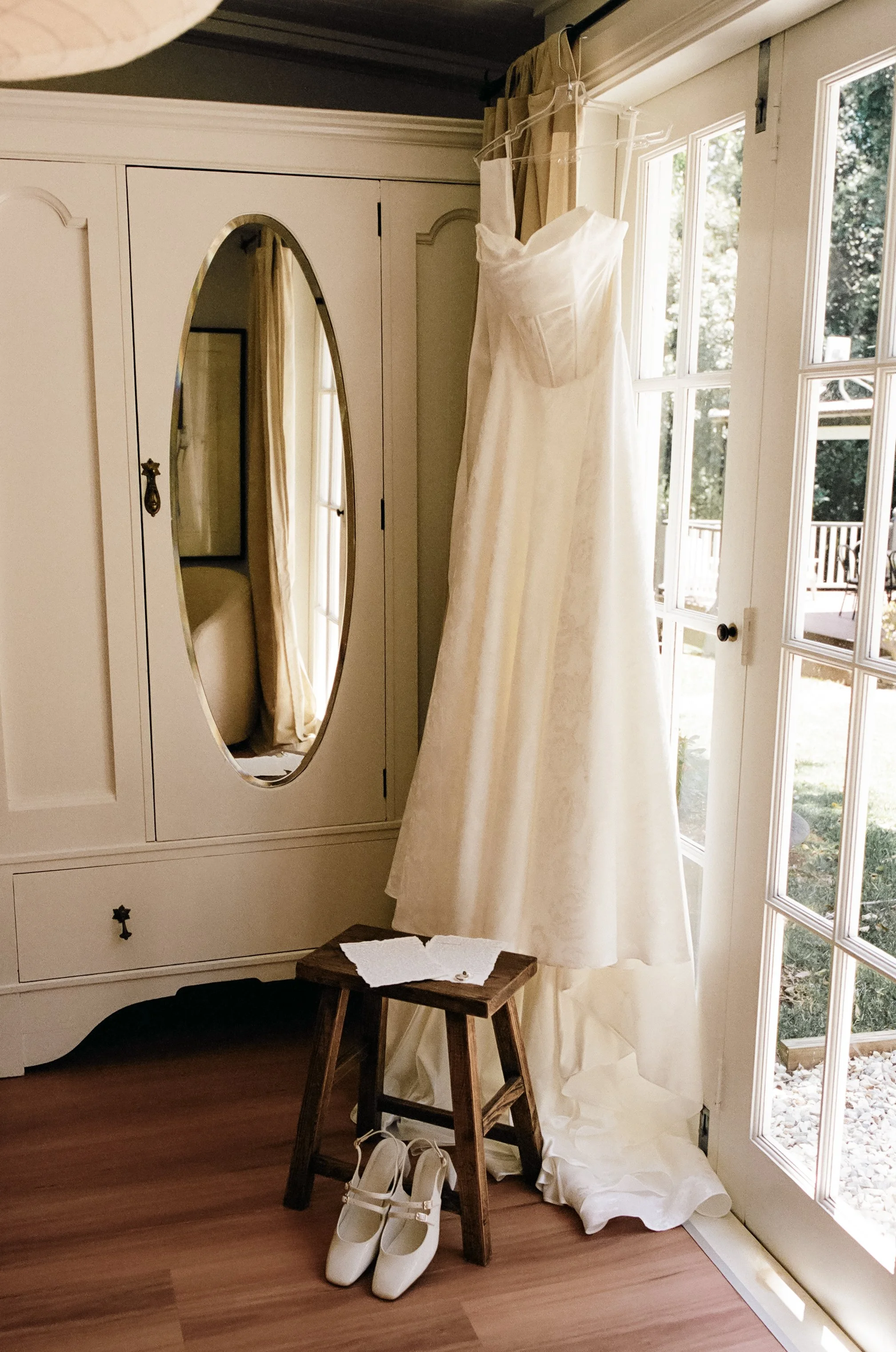 Wedding dress hanging from a hook on a curtain rod near a glass door, with white shoes, a small wooden stool, and notes on top of the stool in a room with beige curtains and a white wardrobe.