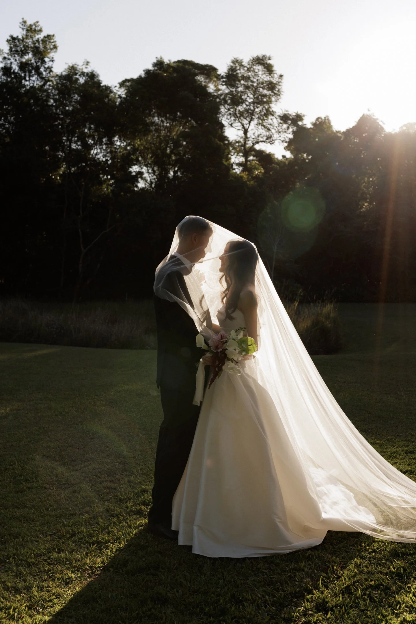A bride and groom standing close together outdoors during sunset, with the bride wearing a white wedding dress and veil, holding a bouquet, and the groom in a black tuxedo. They are facing each other, smiling, with trees and the setting sun in the ba