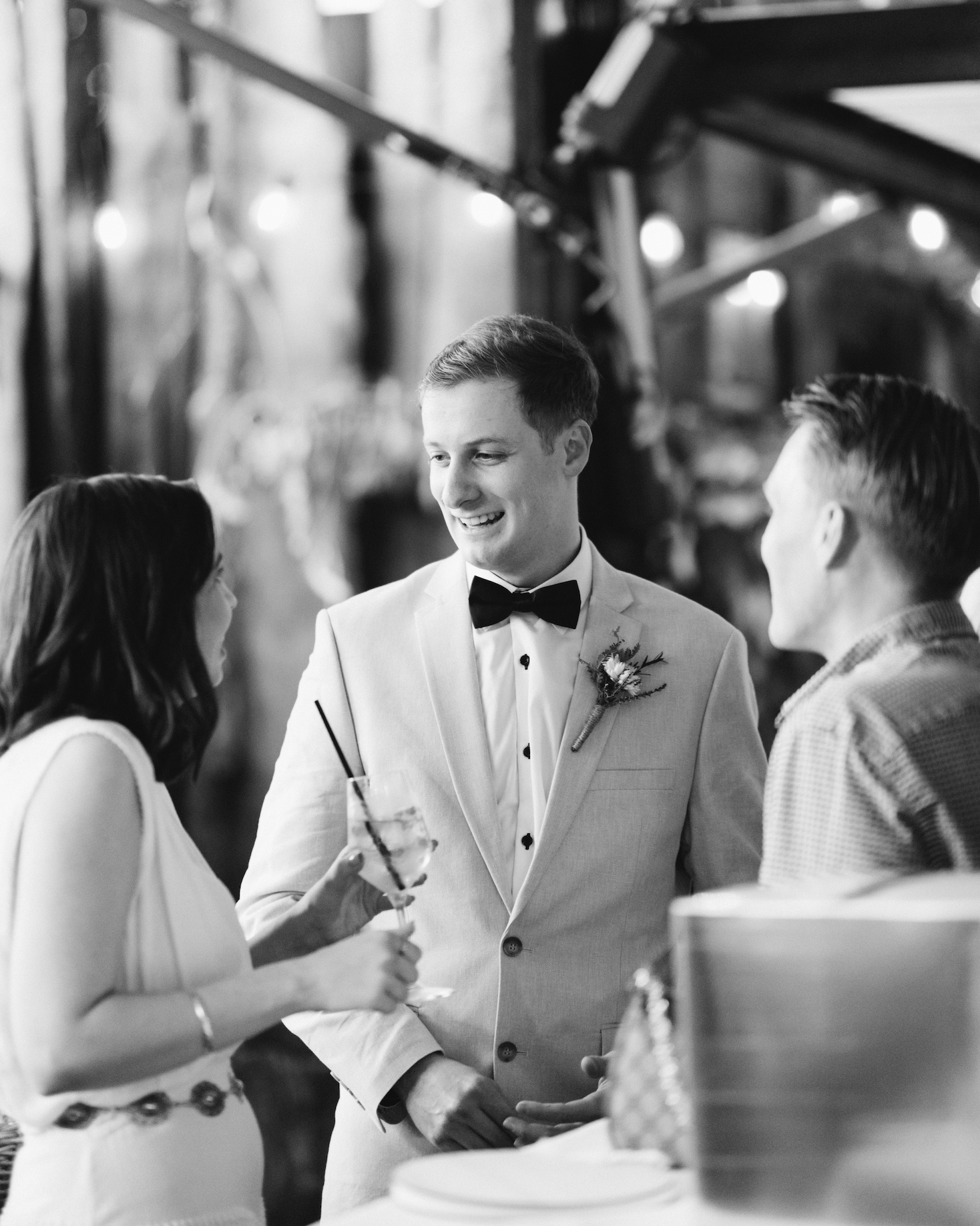 Black and white photo of a man in a tuxedo with a boutonniere talking to a woman holding a drink, with another man listening, at a social event or celebration.