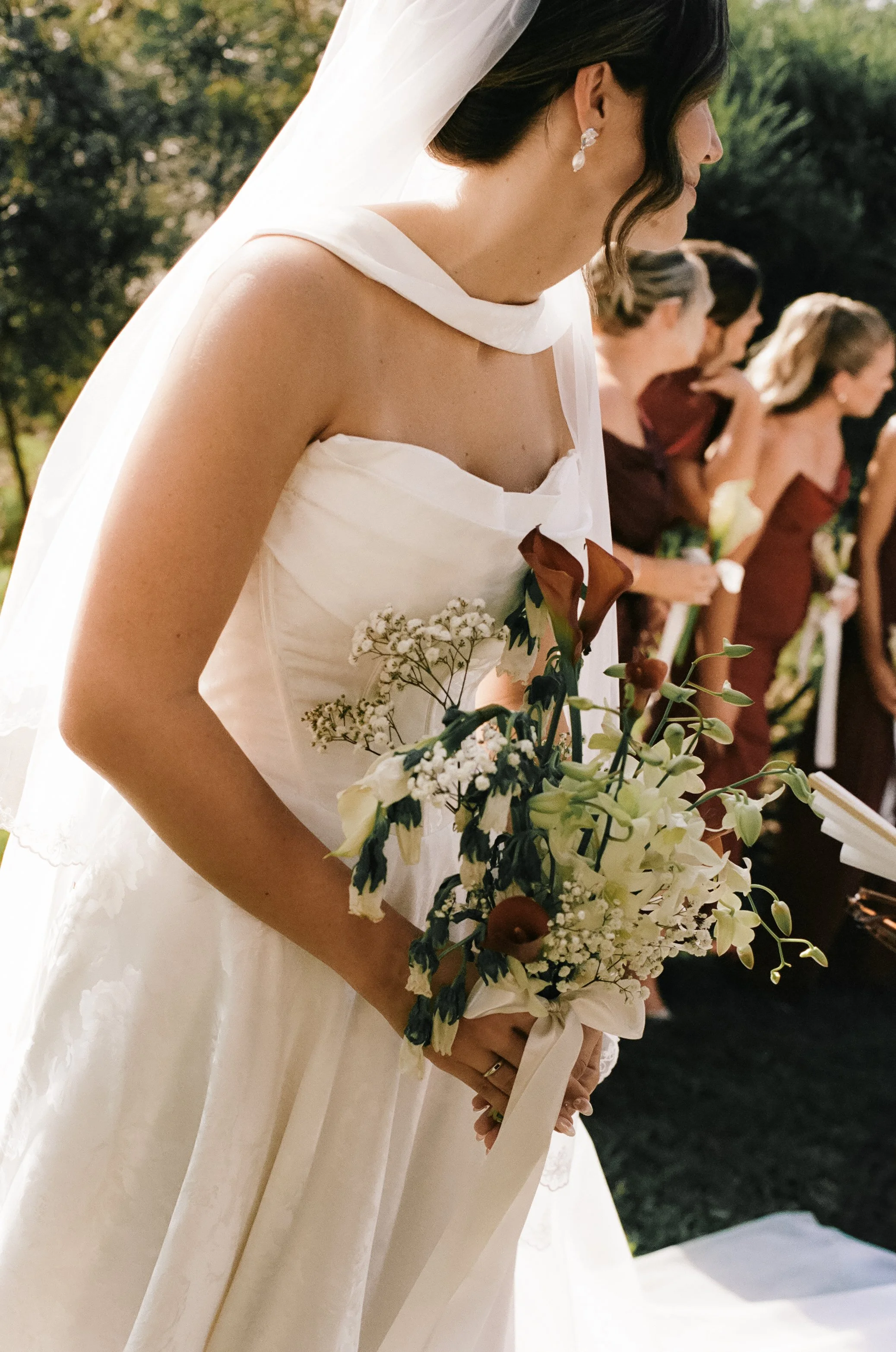 A bride in a white wedding dress holding a bouquet of flowers during a wedding ceremony, with bridesmaids in the background.
