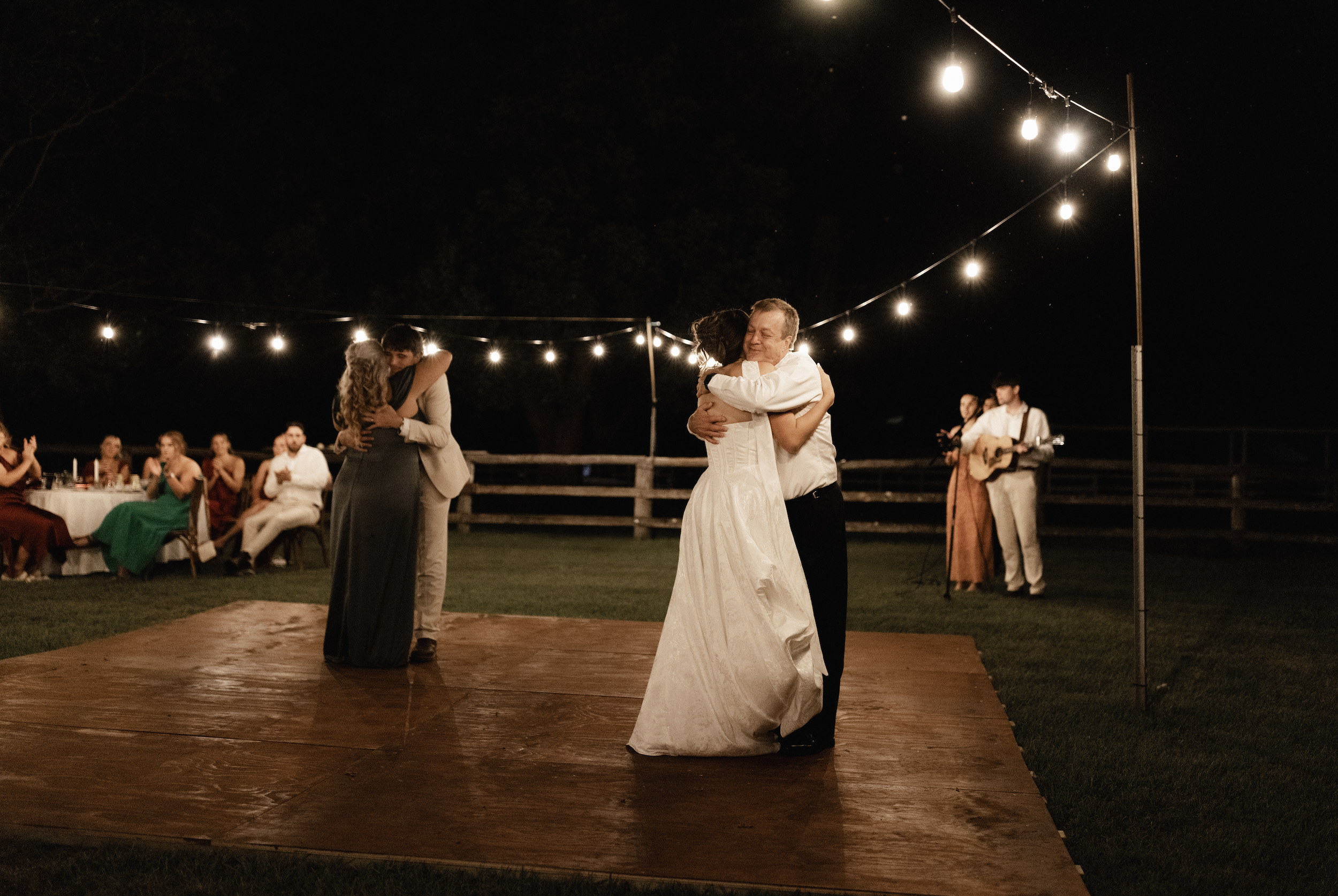 A couple is dancing closely on a wooden platform at an outdoor wedding reception at night. The bride is wearing a white wedding dress, and the groom is in a white shirt with black pants. In the background, another couple is dancing, and guests are se