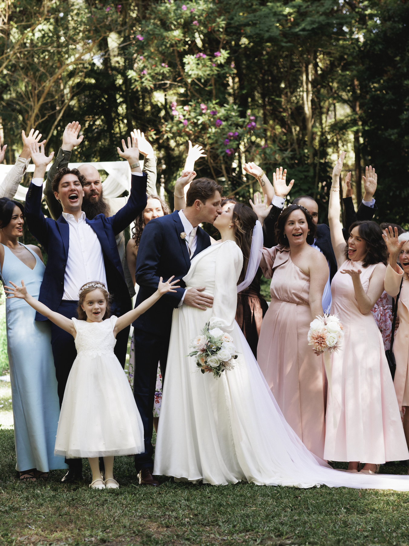 A newlywed couple kissing surrounded by friends and family celebrating outdoors at a wedding.