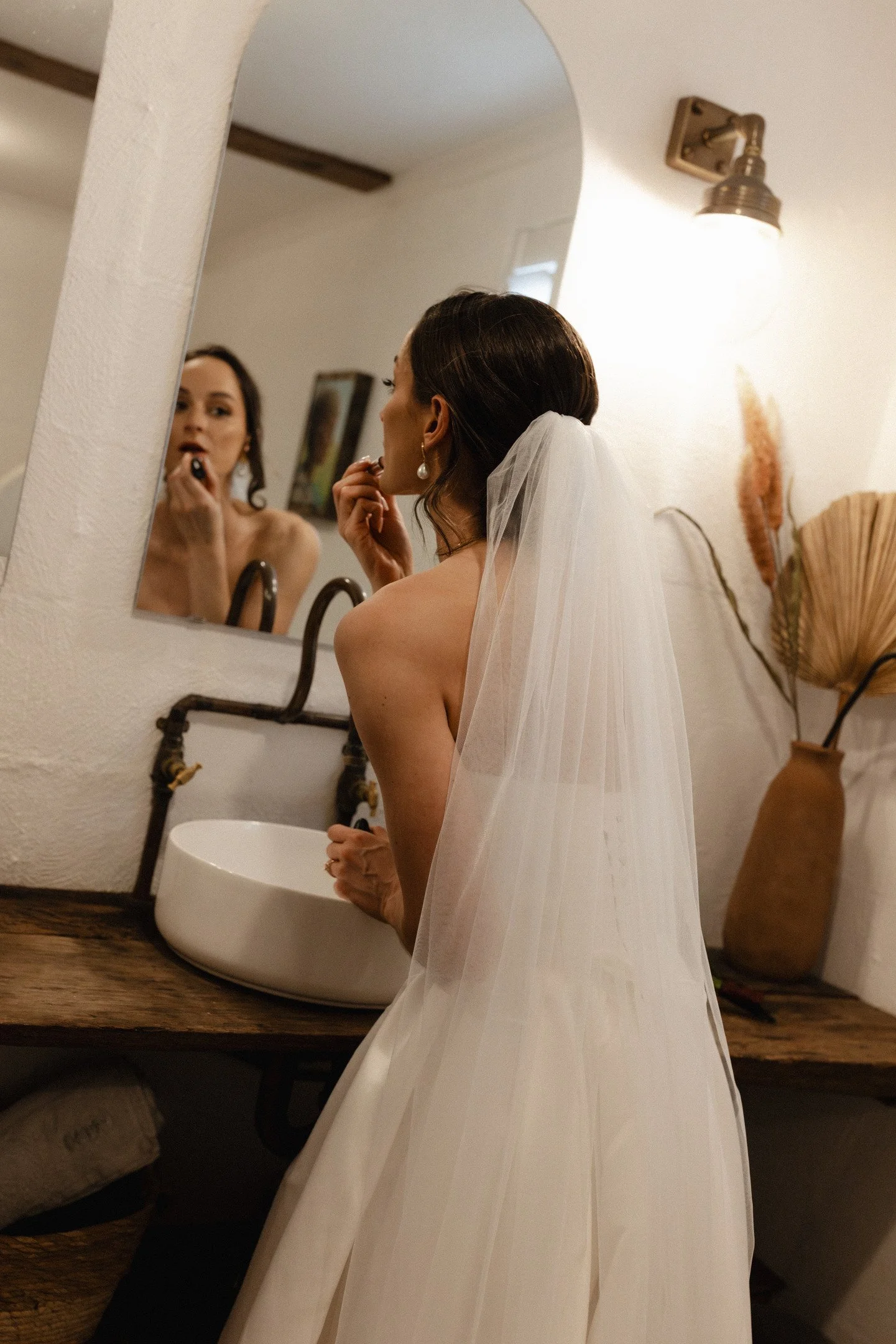 A woman in a white wedding dress with earrings is seen looking into a mirror, applying makeup in a bathroom with warm ambient lighting.