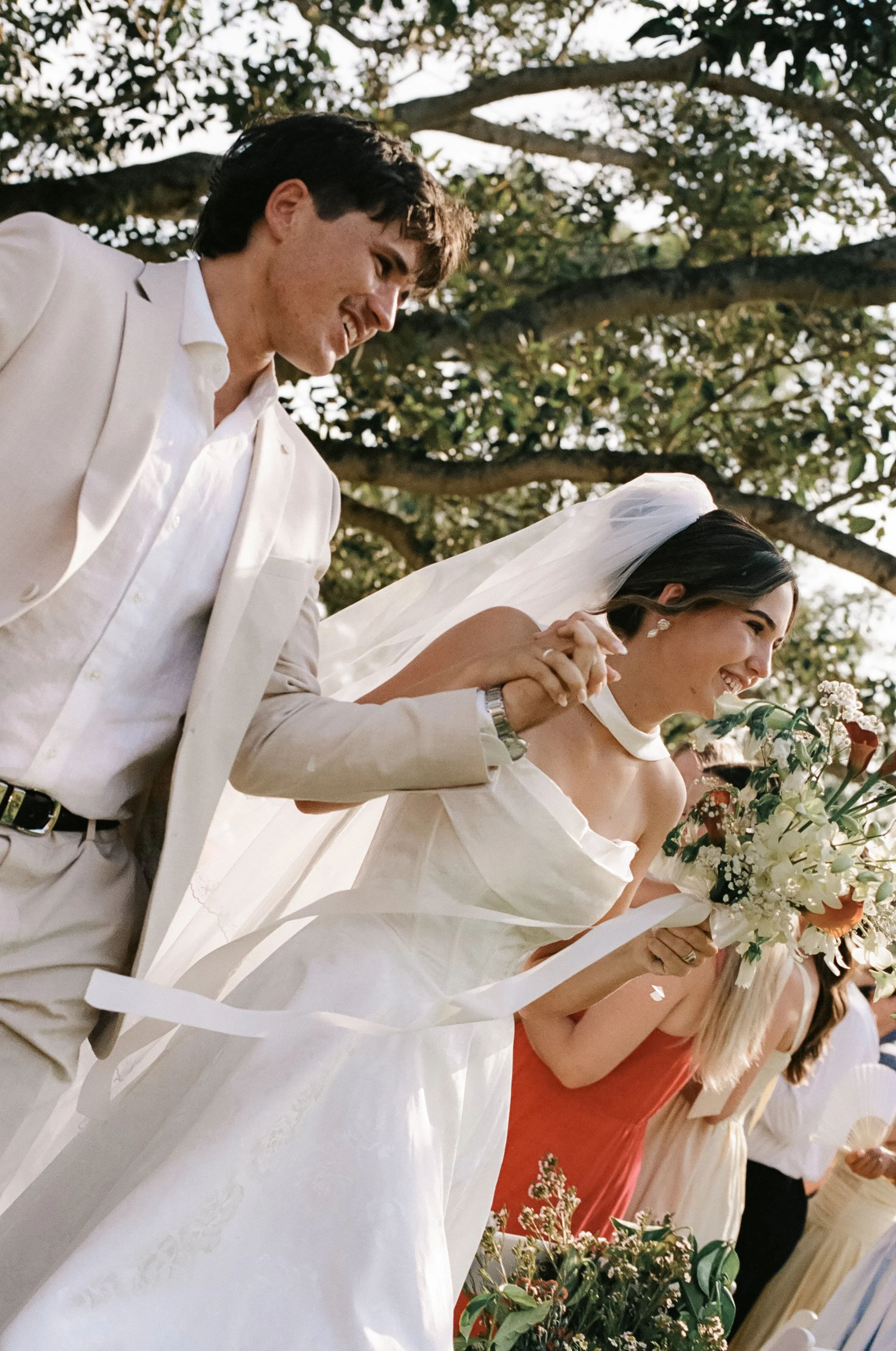 A bride and groom holding hands and smiling during their wedding ceremony outdoors, with guests in the background.
