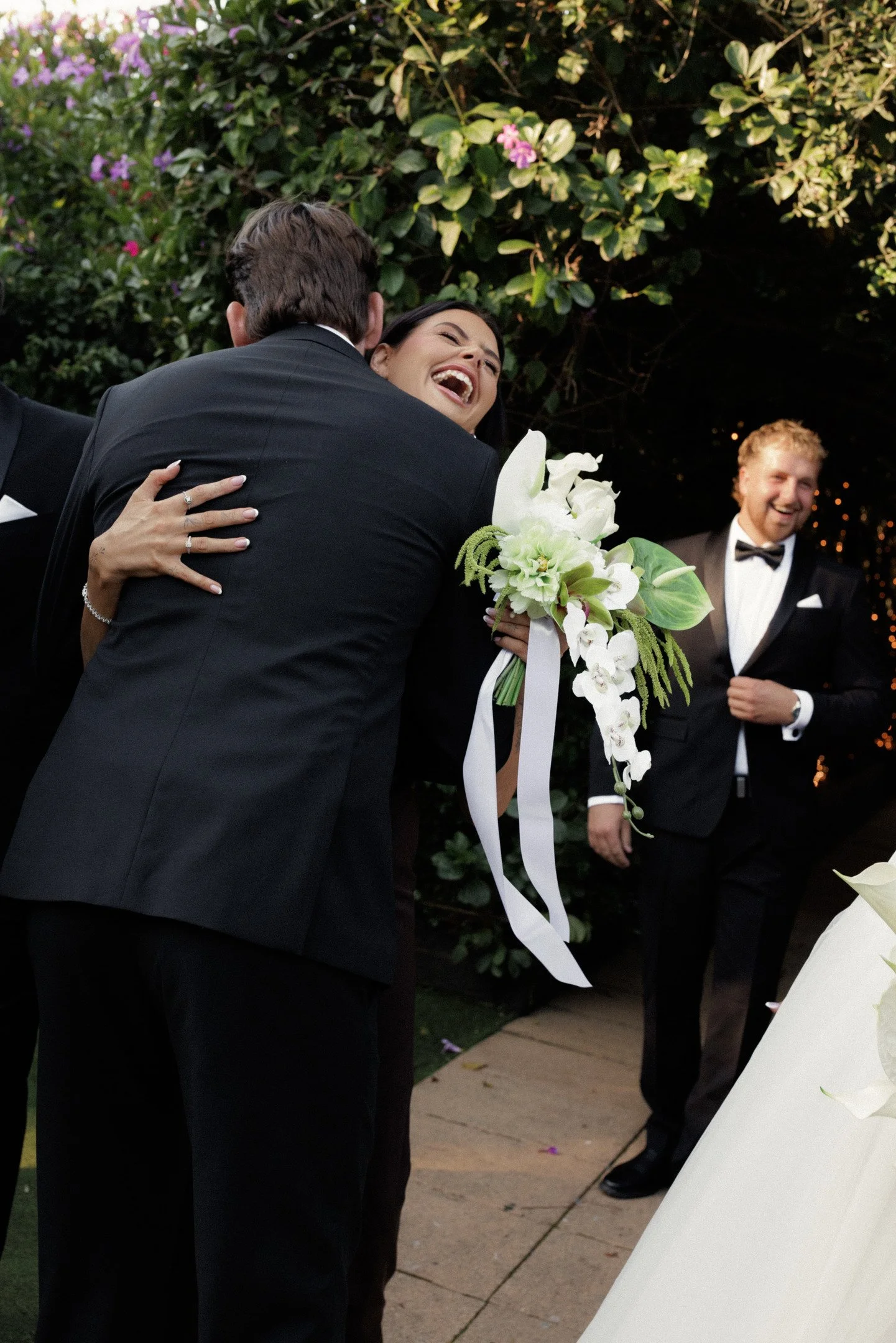 A woman in a black dress and man in a tuxedo hugging at an outdoor wedding reception, with another man in a tuxedo smiling in the background, surrounded by greenery and flowers.
