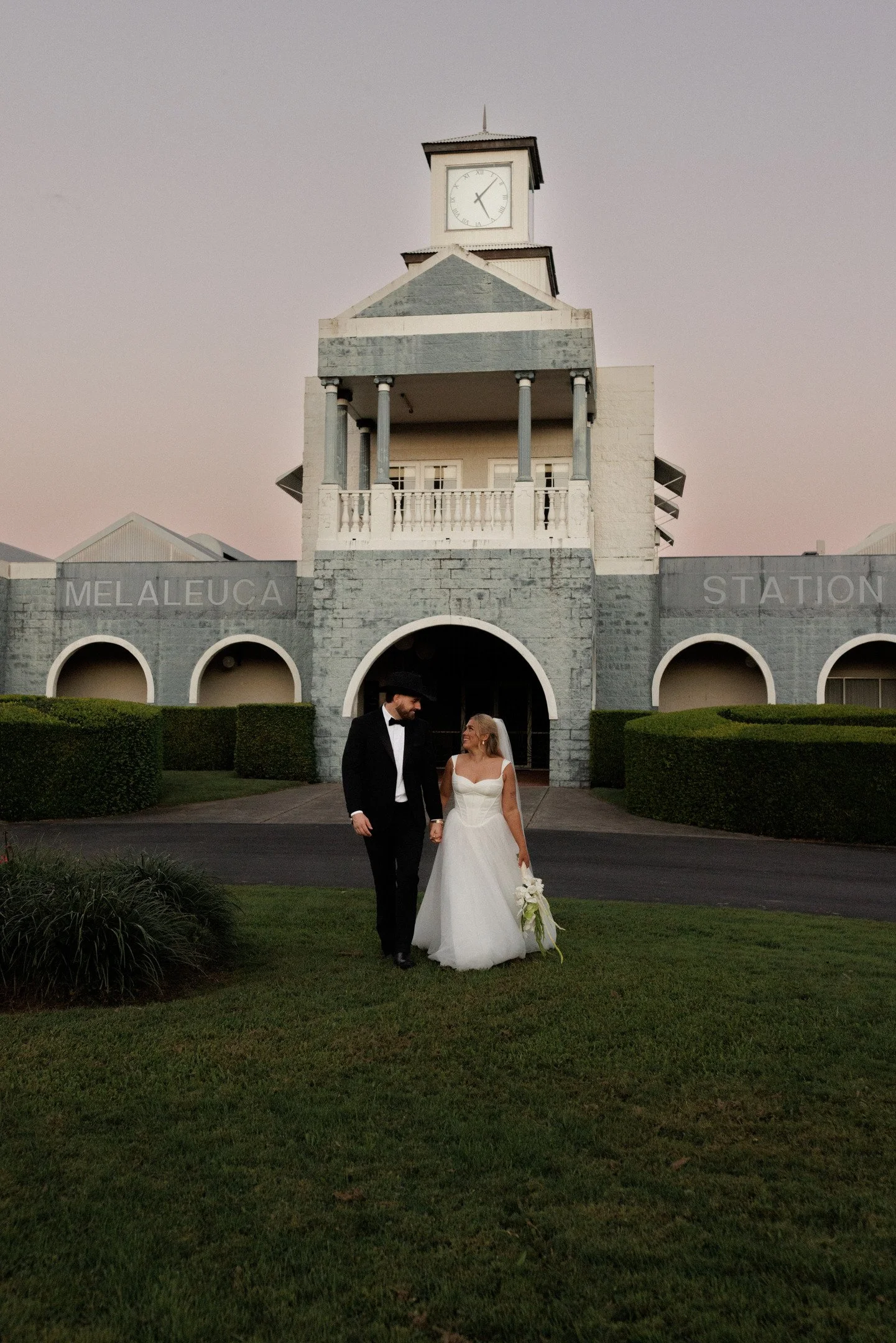 A bride and groom holding hands and walking on a well-manicured lawn in front of a gray stone building with arches and a clock tower at dusk.