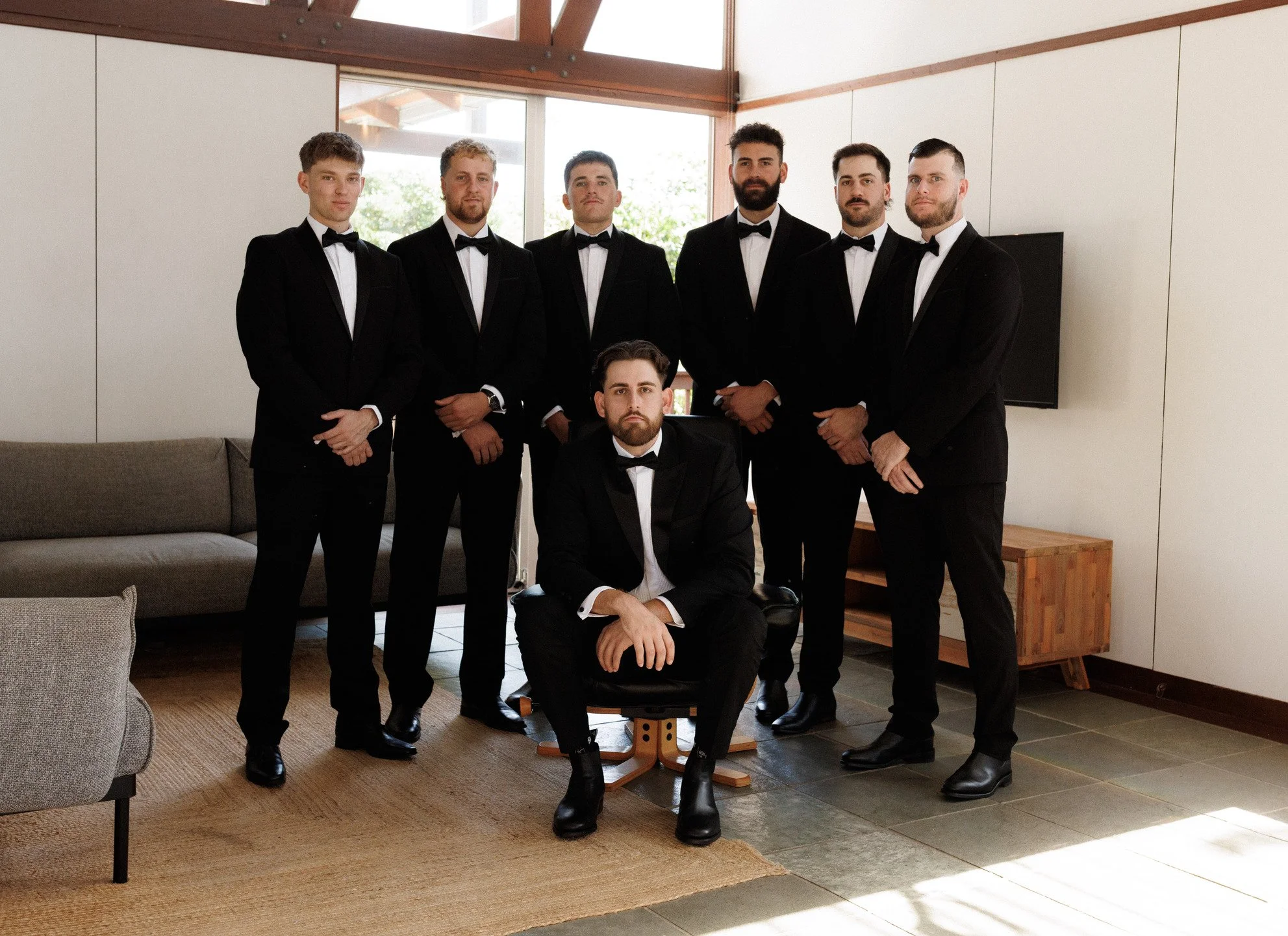 A group of seven men in black tuxedos with bow ties posing in a room with large windows and natural lighting, with one man sitting in front on a chair and six men standing behind him.