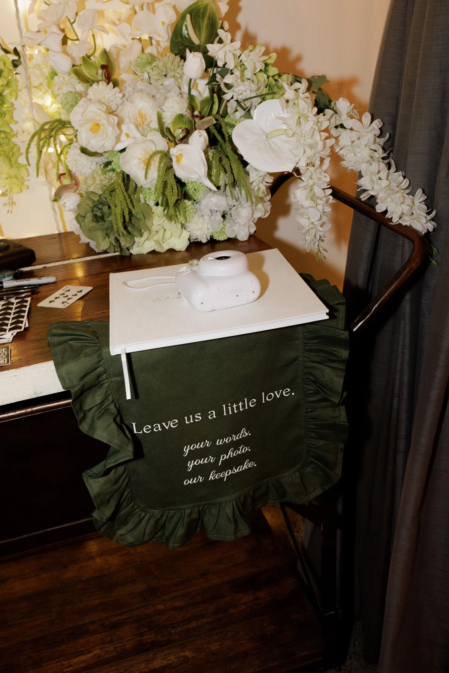A floral arrangement of white flowers, including orchids and other blooms, on a wooden table. A white instant camera is placed on a white book on a dark green cloth-covered table with ruffled edges, with a message printed on the cloth that reads: "Le