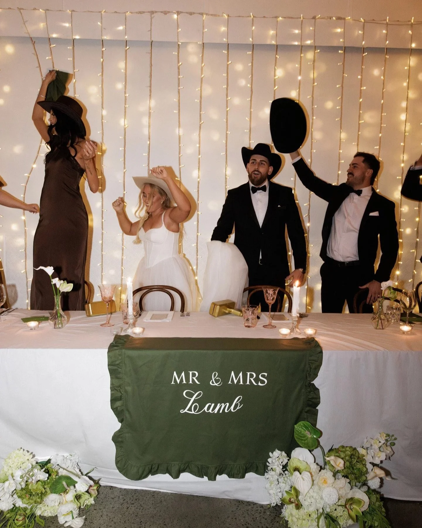 People celebrating at a wedding reception with a table labeled 'Mr & Mrs Lamb' in front of a decorated backdrop of string lights. Guests are wearing formal attire, including hats, with a man holding a black hat in the air.