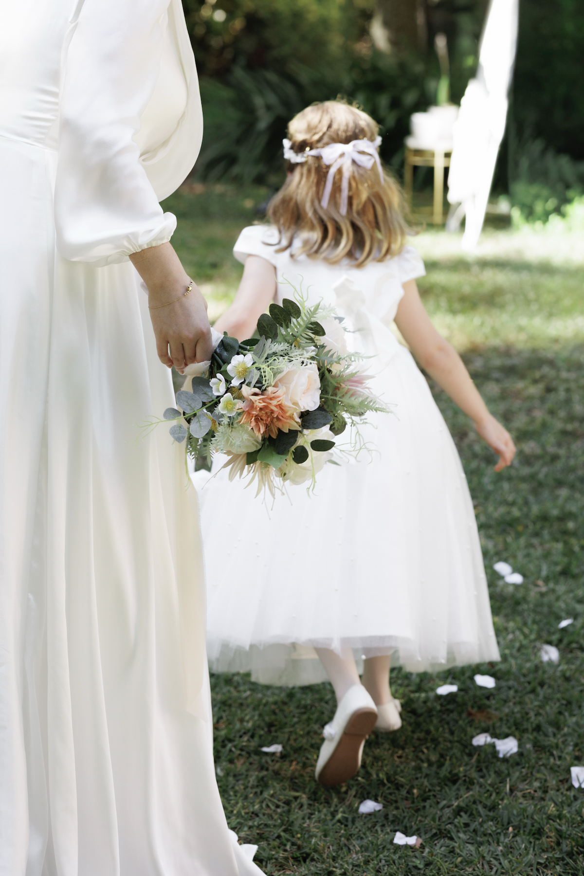 A woman and a young girl in white dresses at an outdoor wedding, with the woman holding a bouquet of flowers and the girl walking on grass with white flower petals.