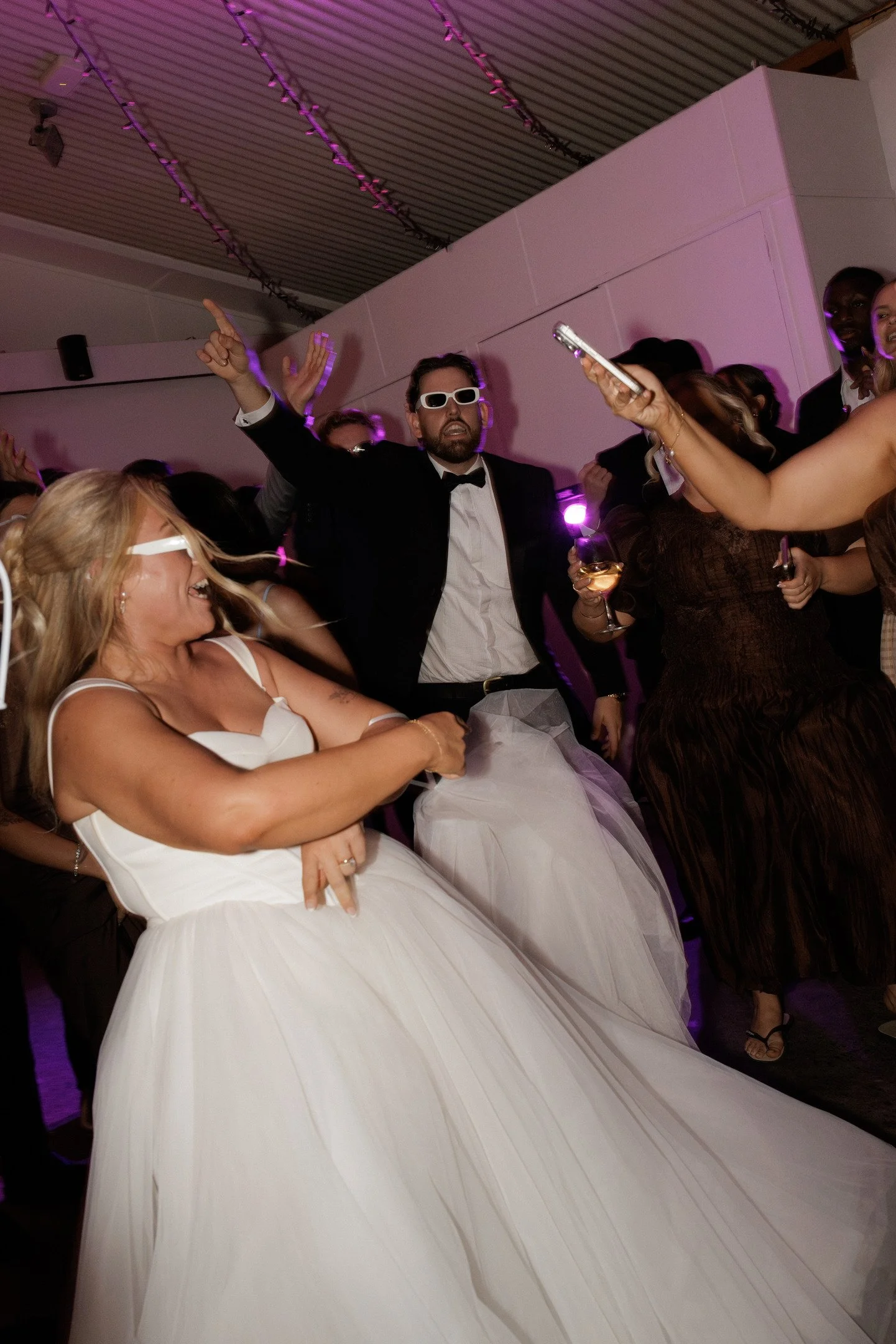 People dancing and partying at an indoor event, including a woman in a white dress, a man in a tuxedo and sunglasses, and other guests in formal attire.
