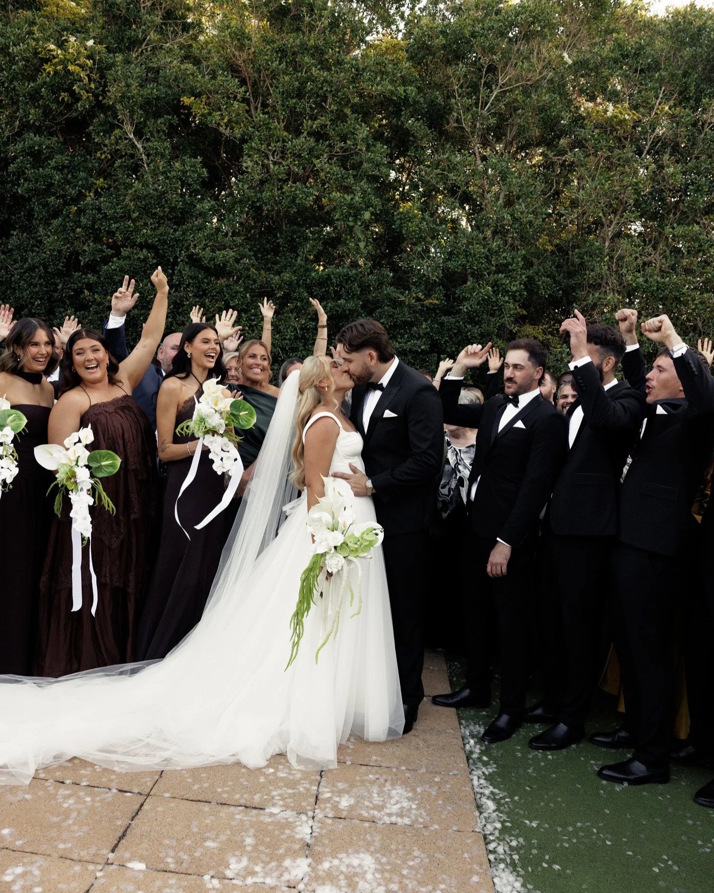A wedding celebration with a bride and groom kissing surrounded by guests dressed in formal attire, outdoors with lush greenery in the background.