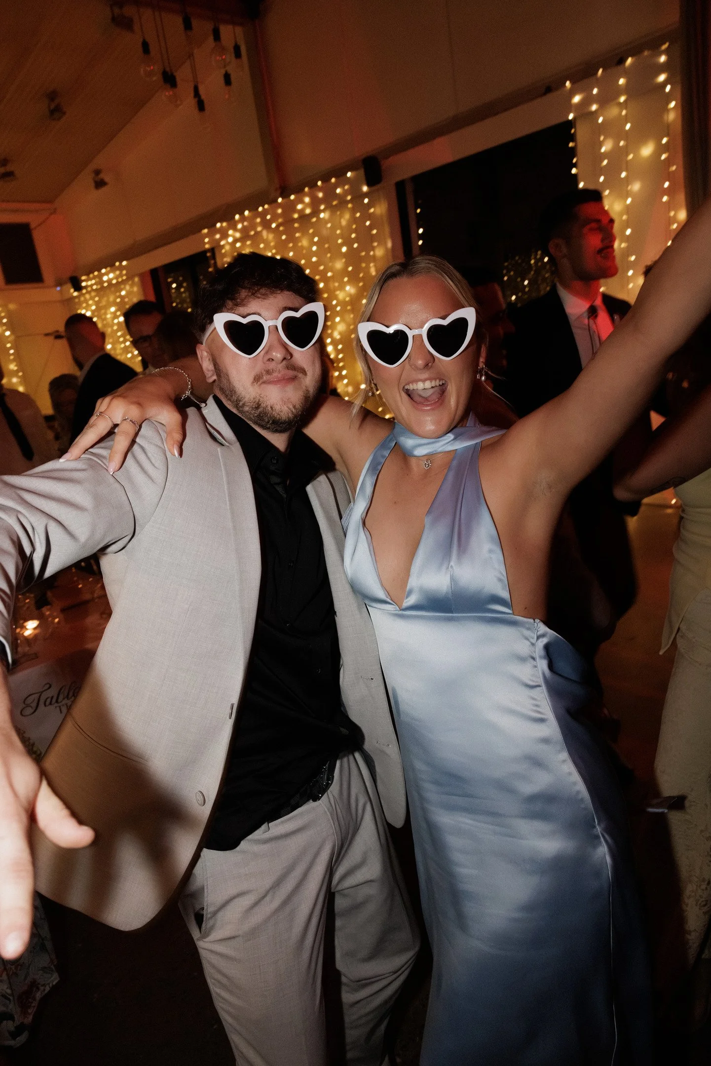 Celebrating party with two people wearing heart-shaped sunglasses and smiling, with string lights in the background.