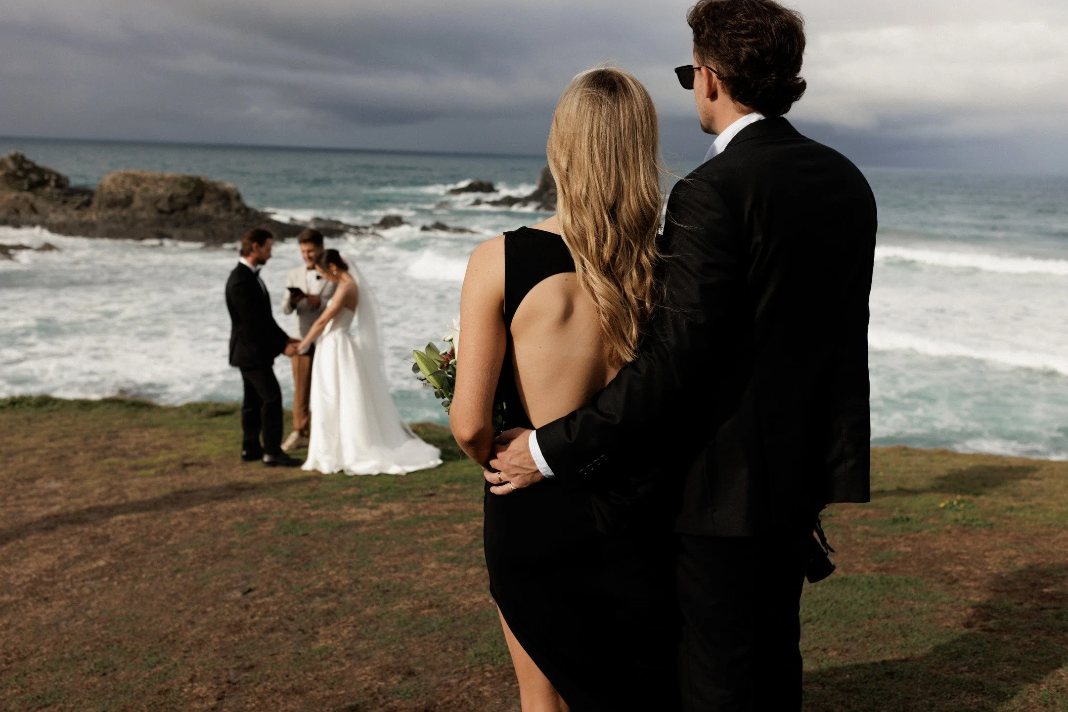 A couple dressed in formal wedding attire stands close together on a grassy area by the ocean, with the groom's arm around the bride's waist. In the background, the bride and groom are exchanging vows with an officiant, and the ocean with rocks and a