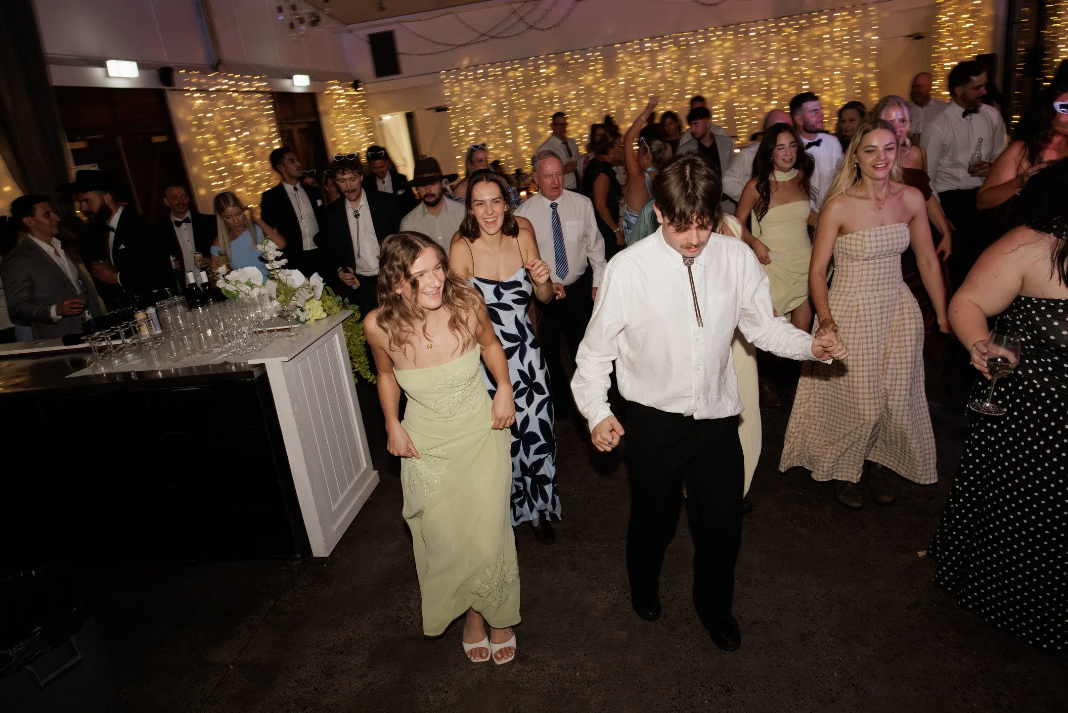 People dancing and enjoying themselves at a celebration with string lights and elegant attire.