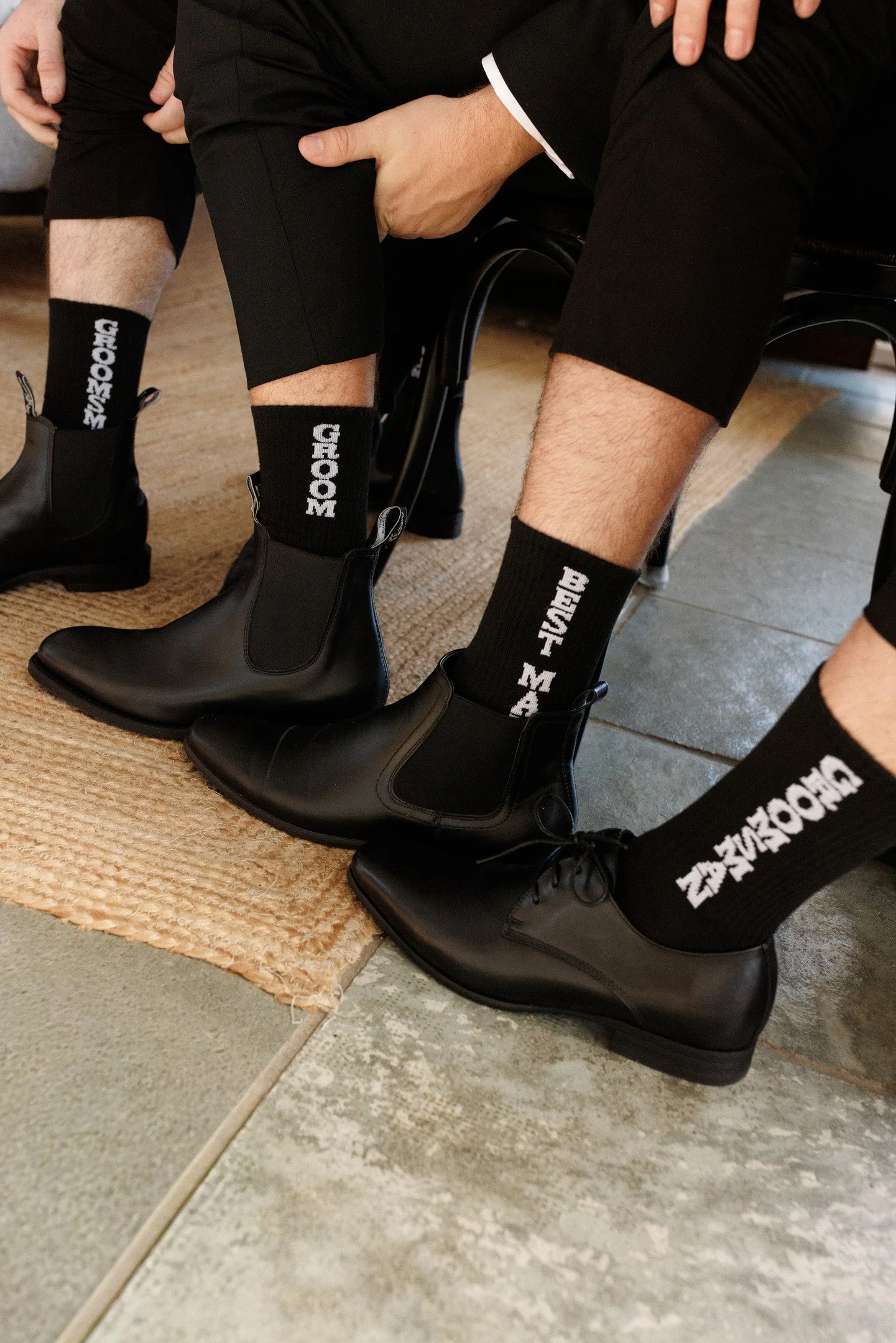 Close-up of two people sitting closely, with one person touching the other's knee. They are wearing black pants, black boots, and black socks with white text that reads "GROOM" and "BRIDE." The scene is indoors, with a beige rug and tiled floor.