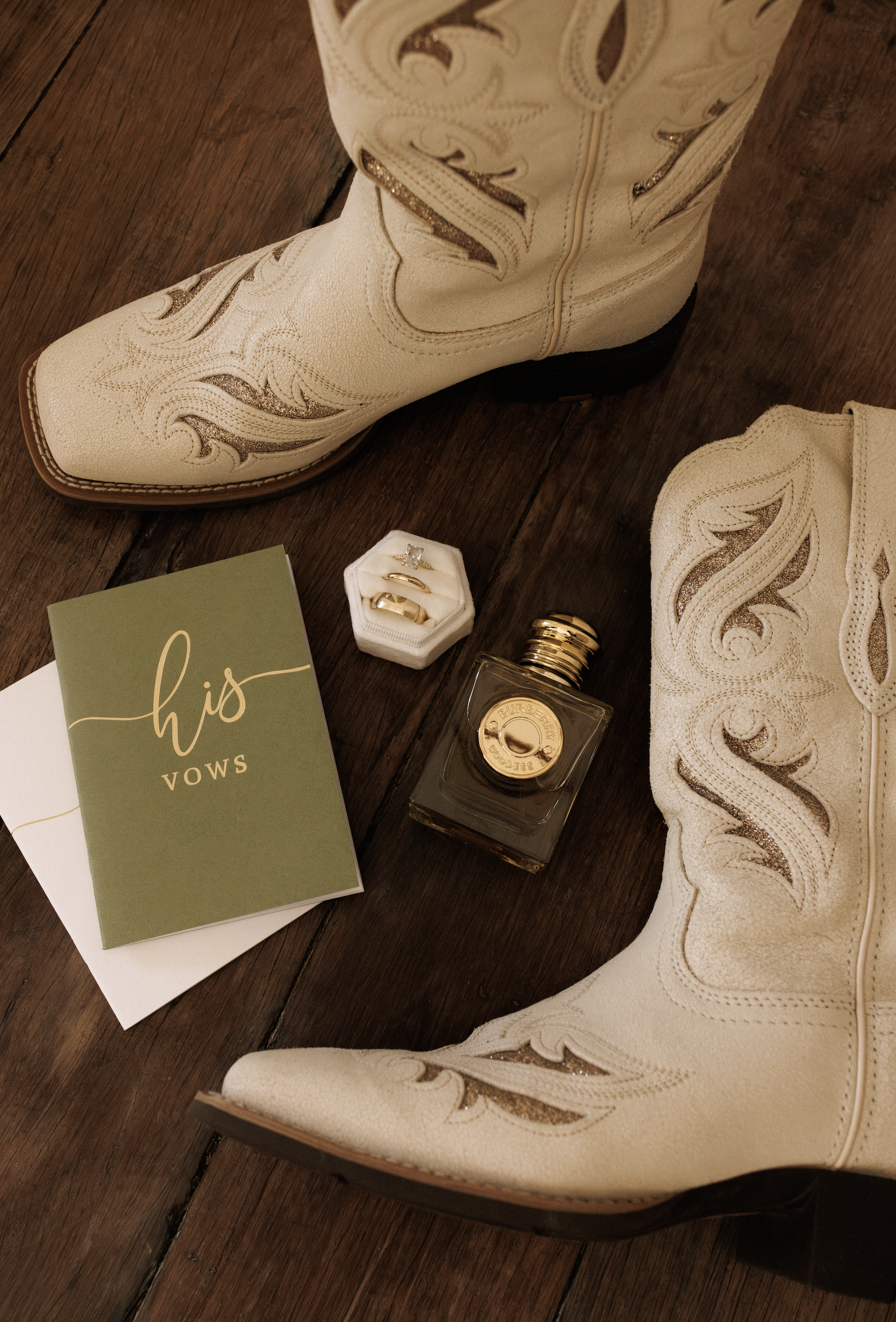 Close-up of white cowboy boots with gold accents, a small perfume bottle, a ring in a white box, and a green card with the word 'his' on a wooden floor.