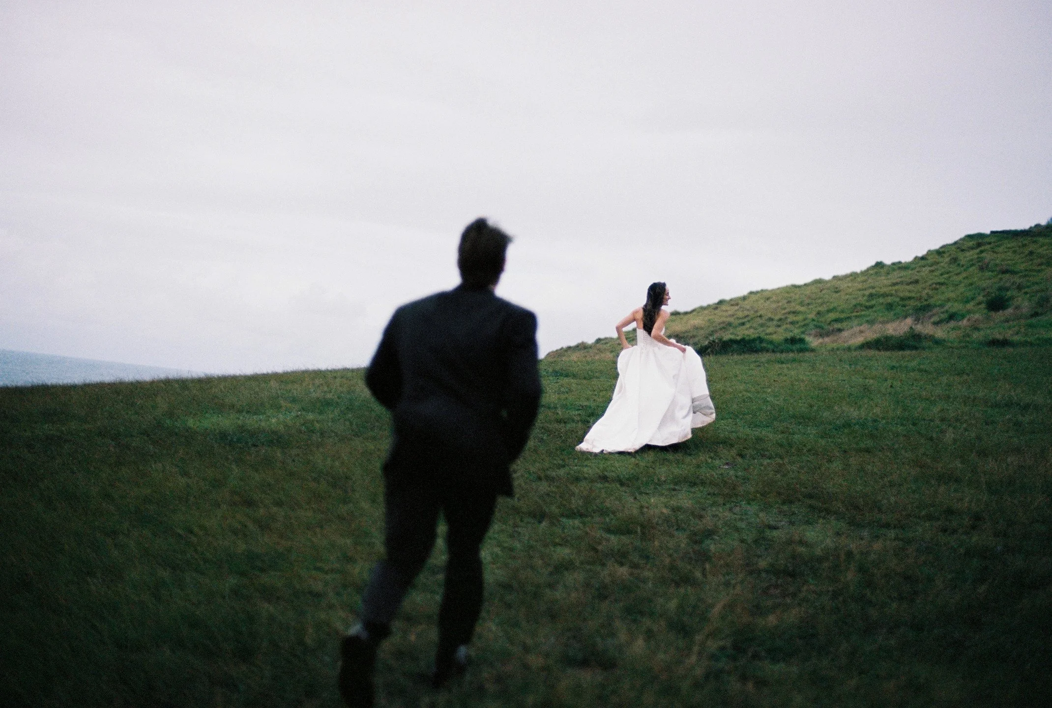 A man and woman in formal attire outside on a grassy hill, with the woman in a white gown and the man in a dark suit, possibly during a photoshoot or event.
