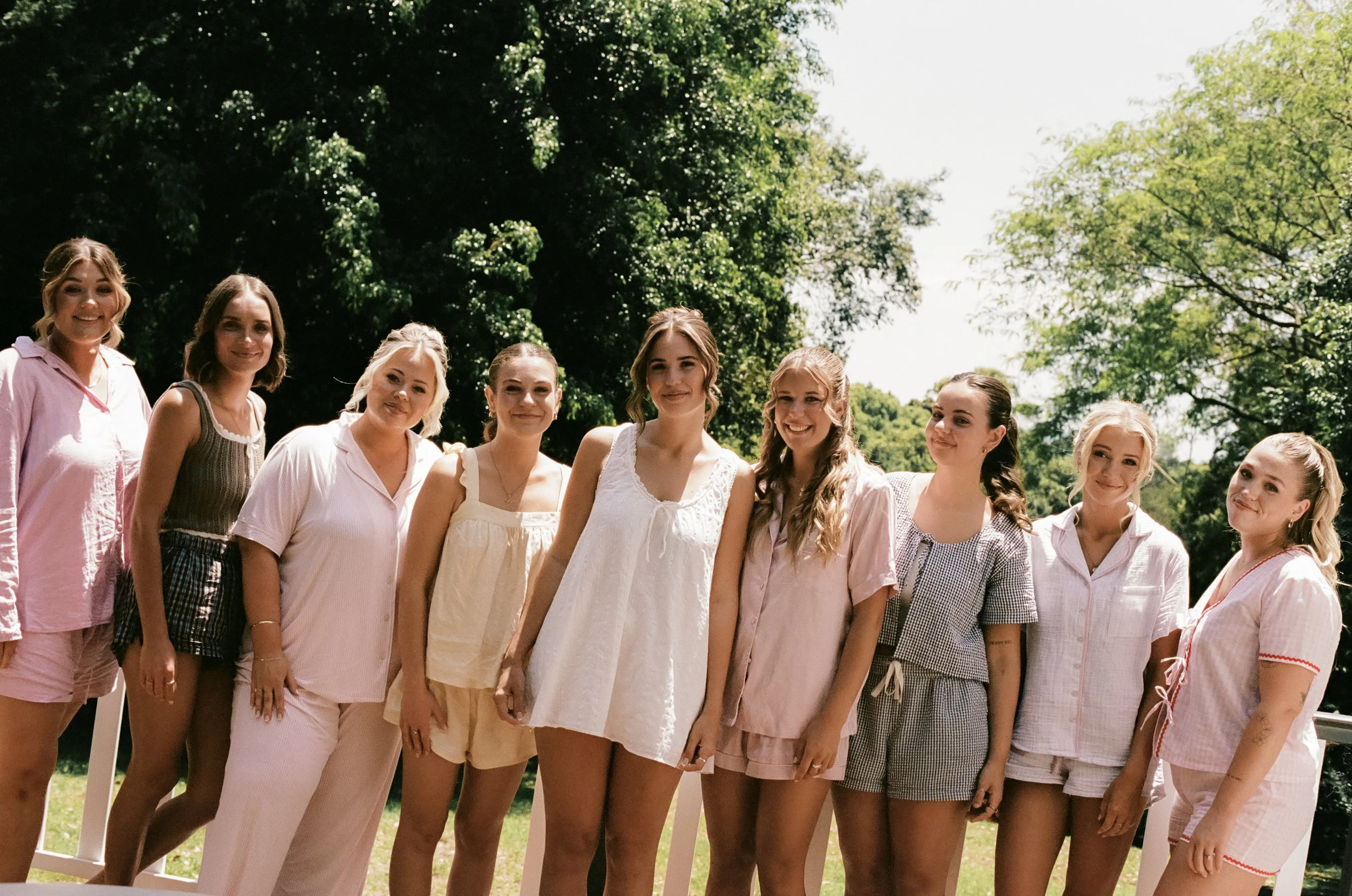 A group of ten young women standing outdoors in pajamas, smiling at the camera on a sunny day with green trees in the background.