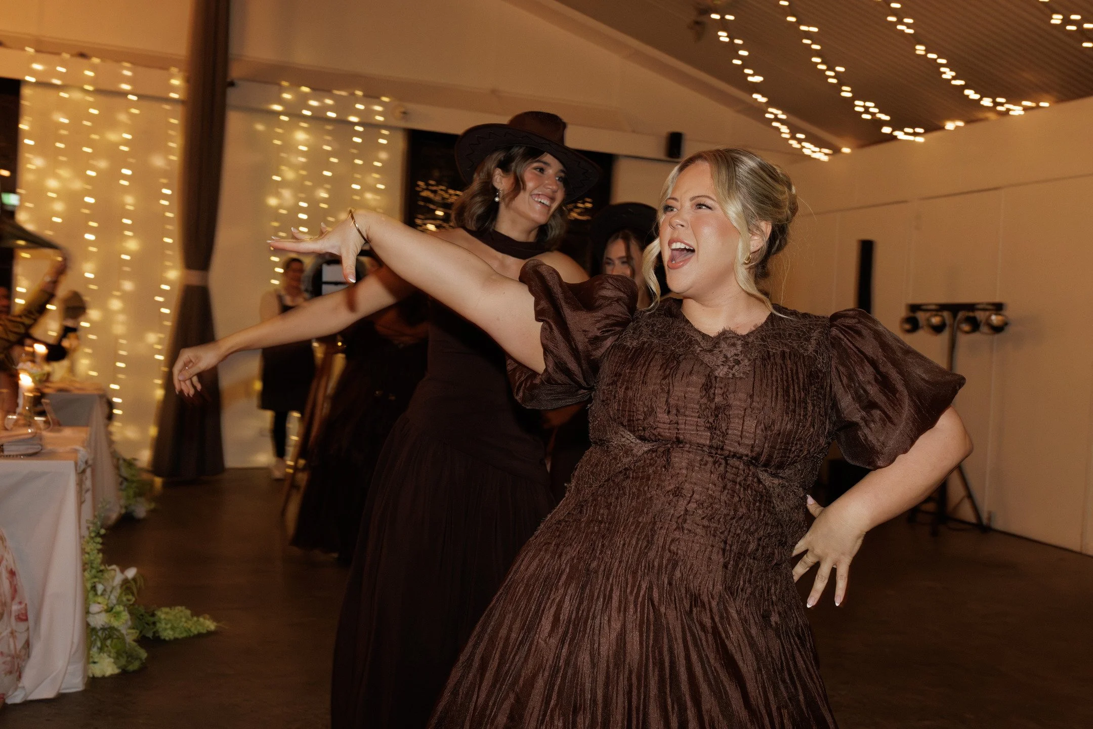 Two women dancing with joyful expressions at an indoor event with string lights in the background.