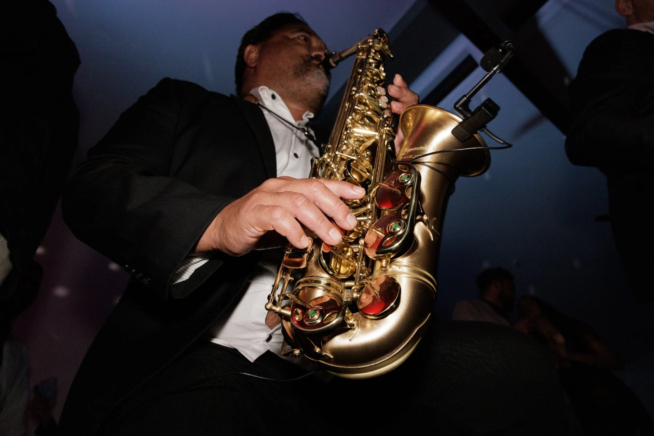 A man playing a gold saxophone, wearing a black suit, white shirt, and a necklace, in a dimly lit setting.