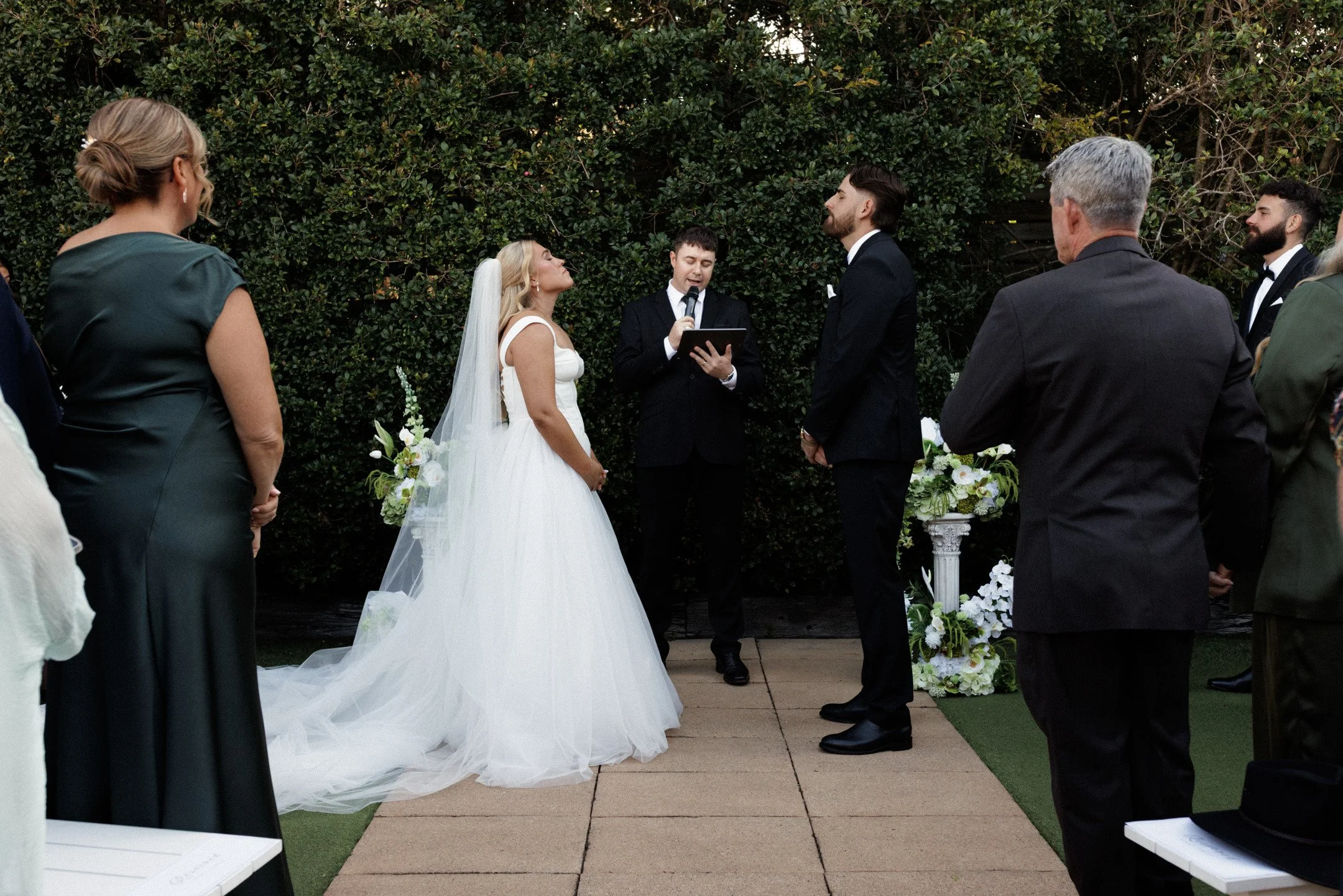 A wedding ceremony outdoors with a bride and groom standing in front of a person officiating. The bride wears a white wedding dress with a veil, and the groom is in a black suit. Guests are gathered around, some facing the couple, with greenery in th