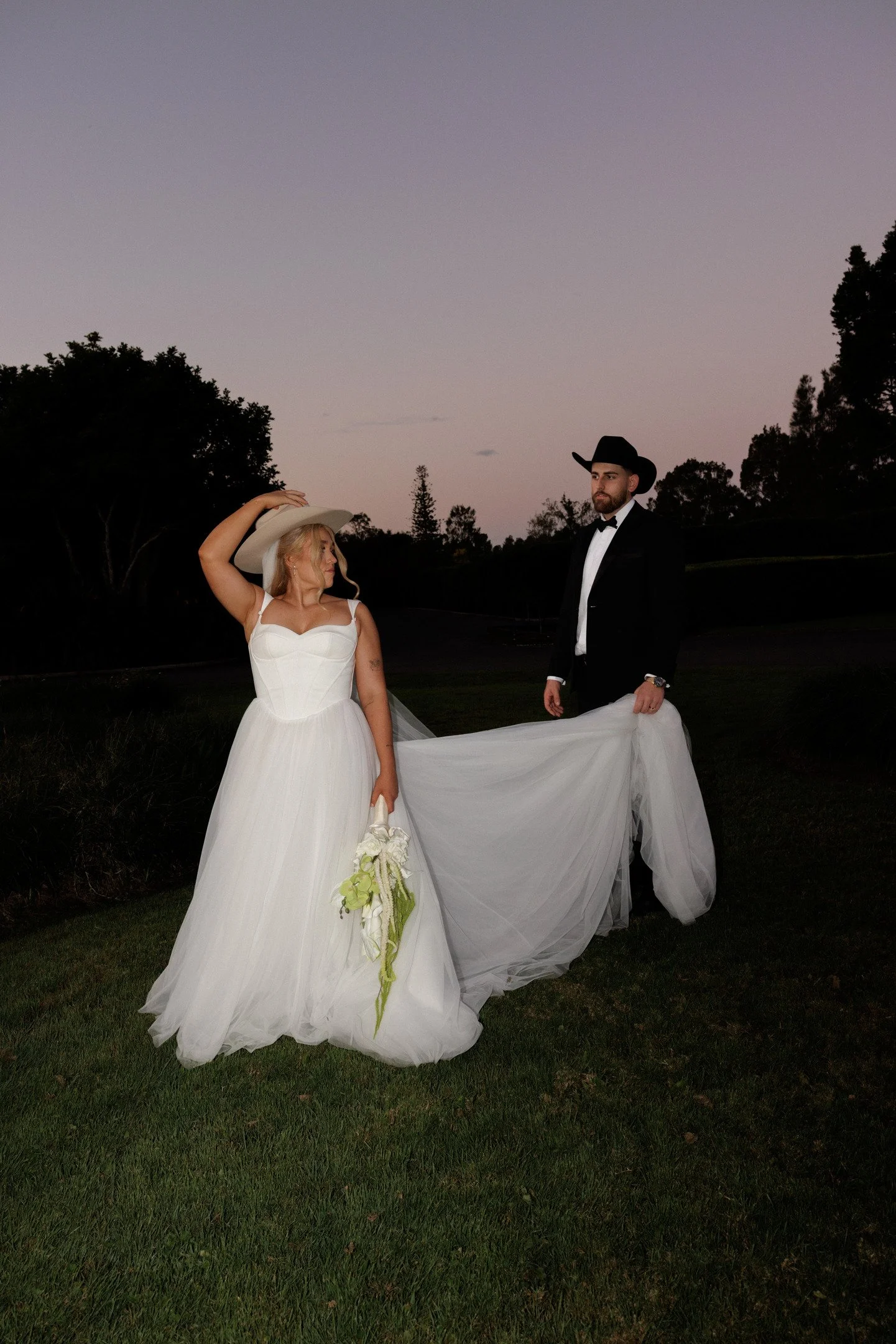 A bride in a white wedding dress and a groom in a black tuxedo with cowboy hat standing outdoors during dusk. The bride is holding a bouquet of white and yellow flowers, and the groom is holding the train of her dress. They are surrounded by trees an