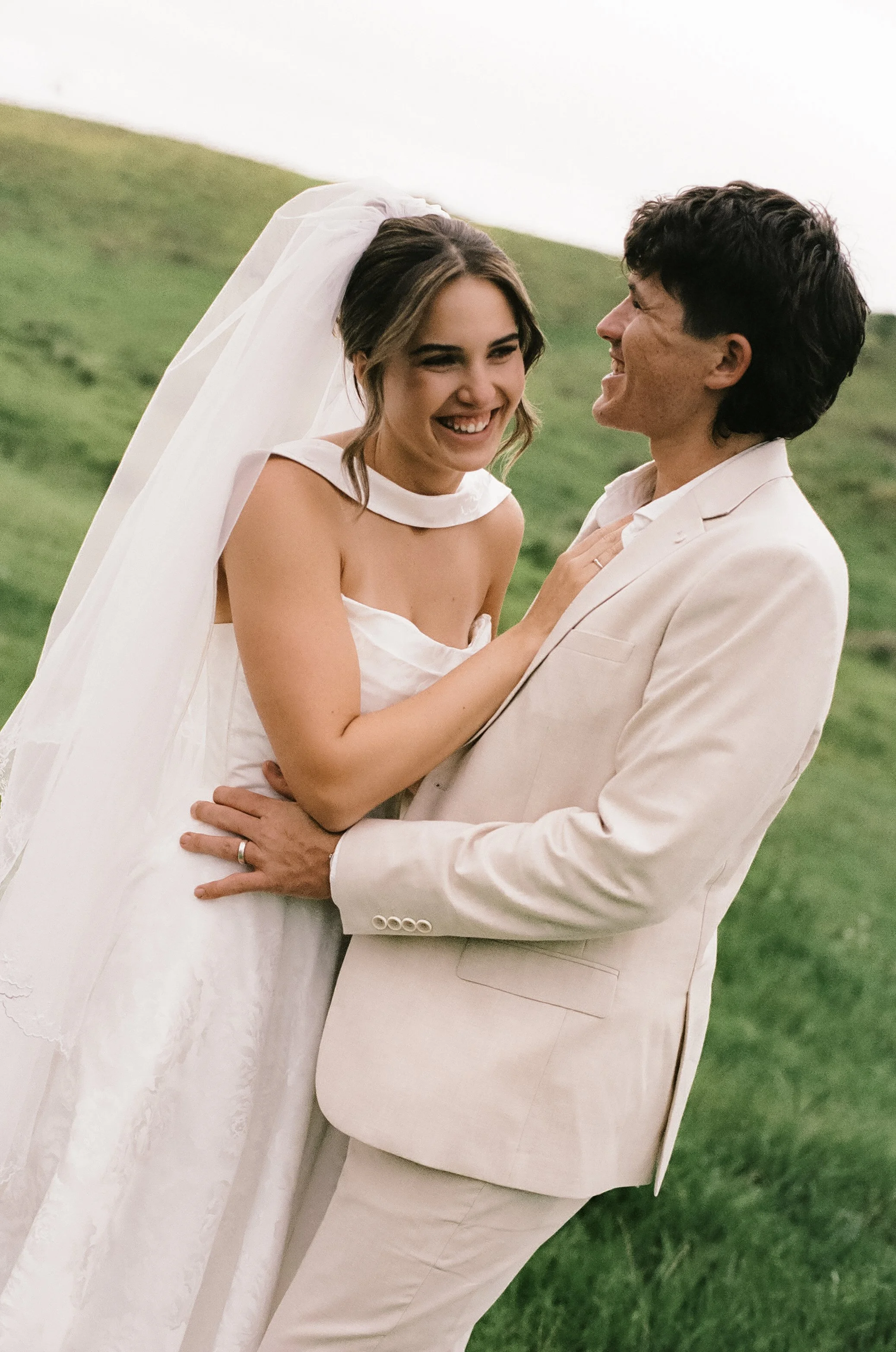 A smiling bride and groom are standing outdoors on a grassy hill, sharing a joyful moment in their wedding attire.