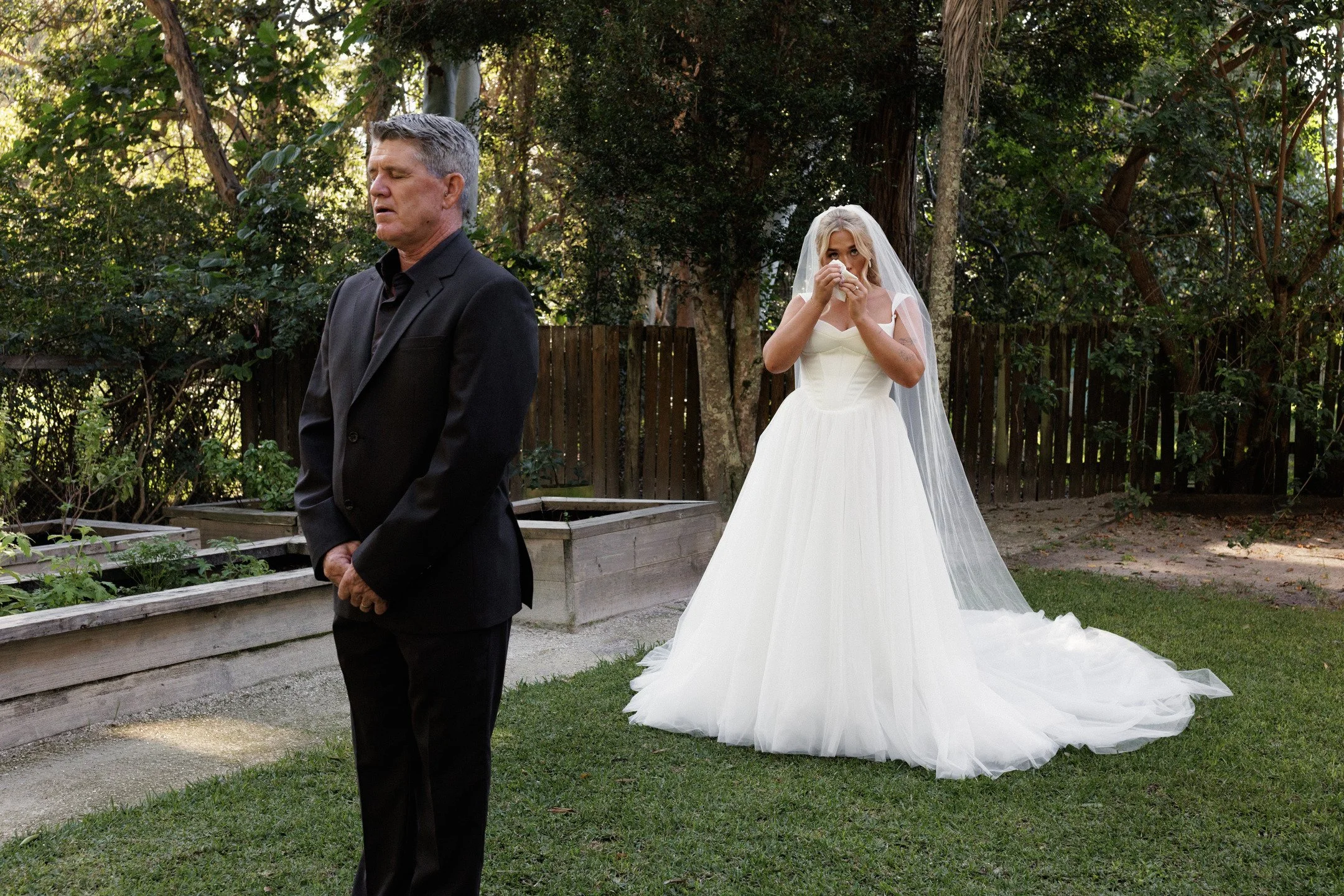 A man in a black suit is standing outside with his eyes closed, while a woman in a white wedding dress and veil appears to be crying and wiping her eyes, set against a background of trees and a wooden fence.