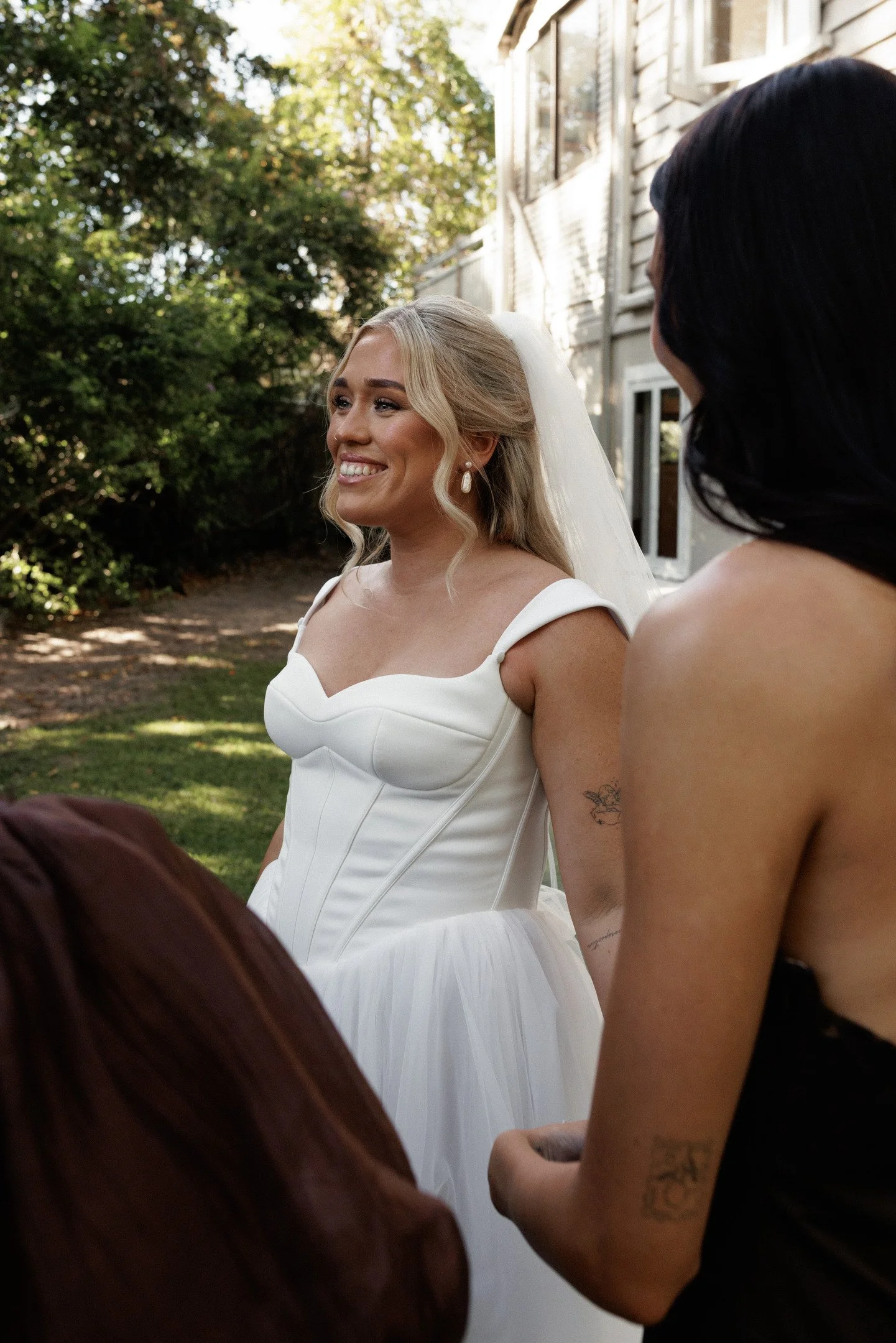 A bride with blonde hair, wearing a white wedding dress and pearl earrings, is smiling while standing outdoors near an older woman with dark hair and tattoos on her arm, in front of a weathered house with trees in the background.