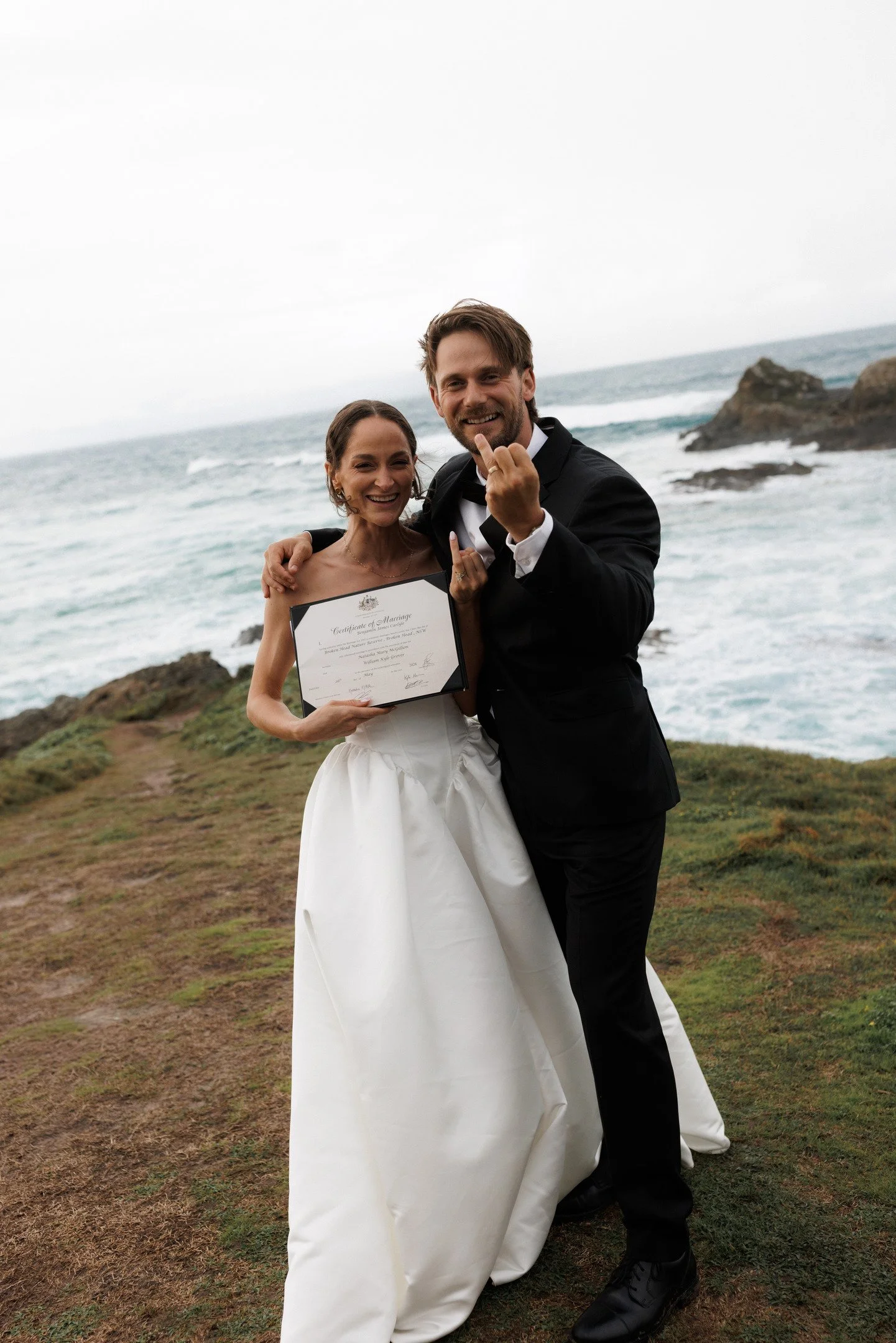A happy couple on their wedding day at the beach, holding a marriage certificate and smiling at the camera. The woman is wearing a white wedding dress, and the man is wearing a black tuxedo. The background features the ocean, rocks, and a cloudy sky.