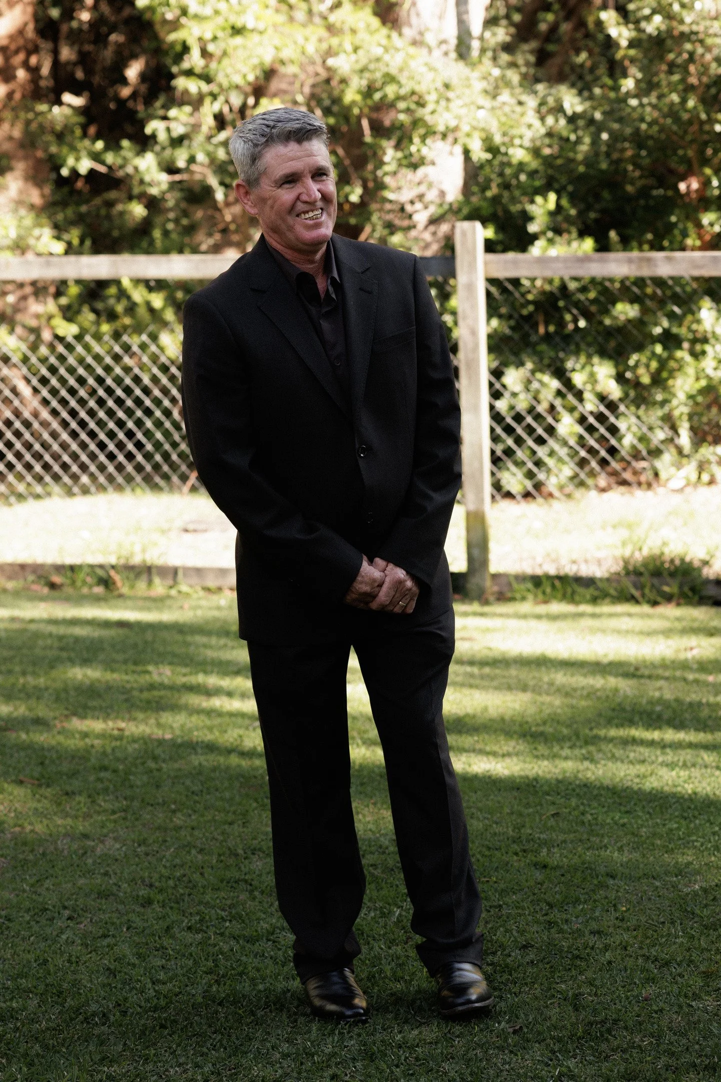 A man in a dark suit smiling outdoors on grass, with a wooden fence and trees in the background.