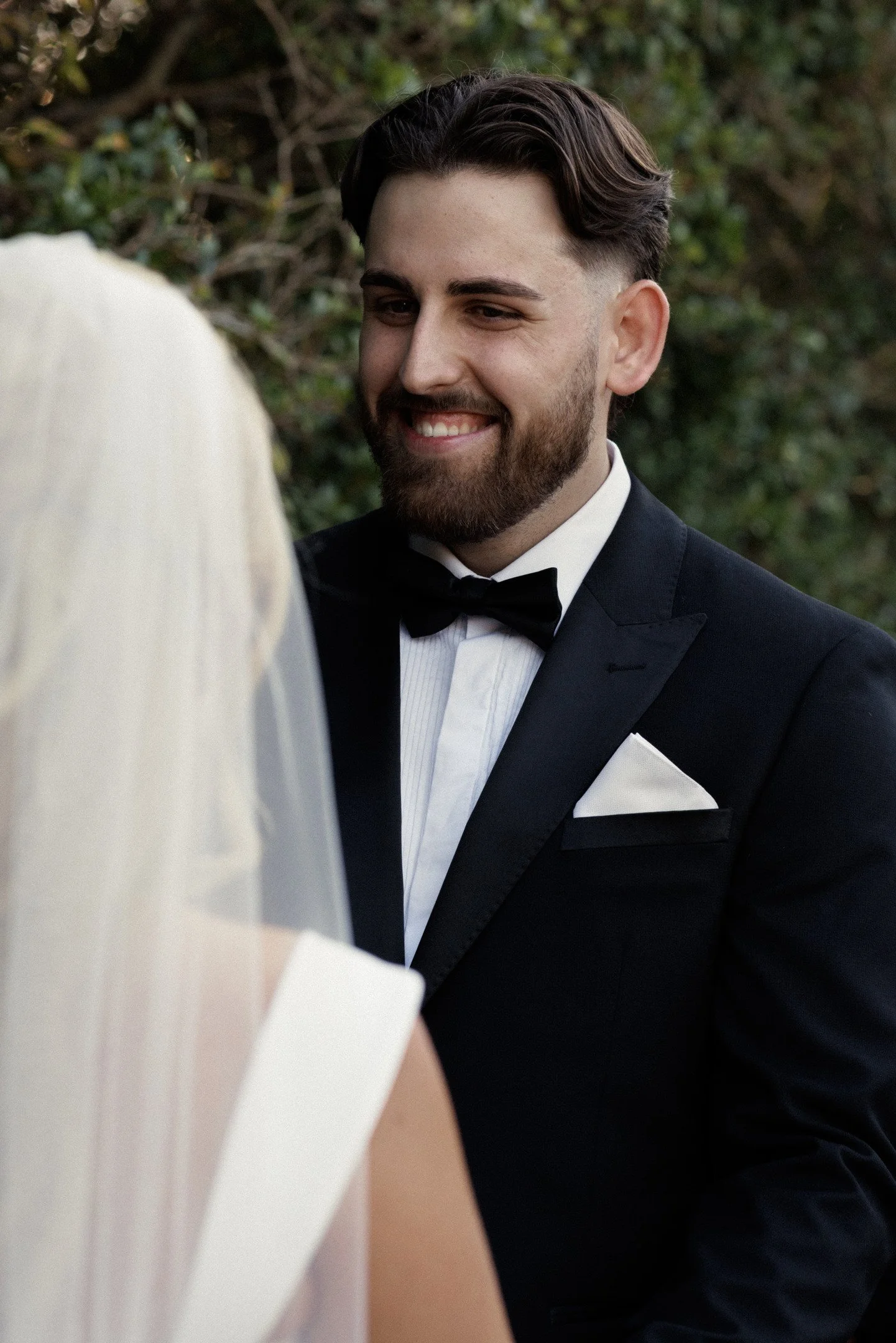 A man dressed in a black tuxedo with a bowtie, smiling during a wedding ceremony, with a woman in a white dress partly visible in the foreground, outdoors with green foliage in the background.