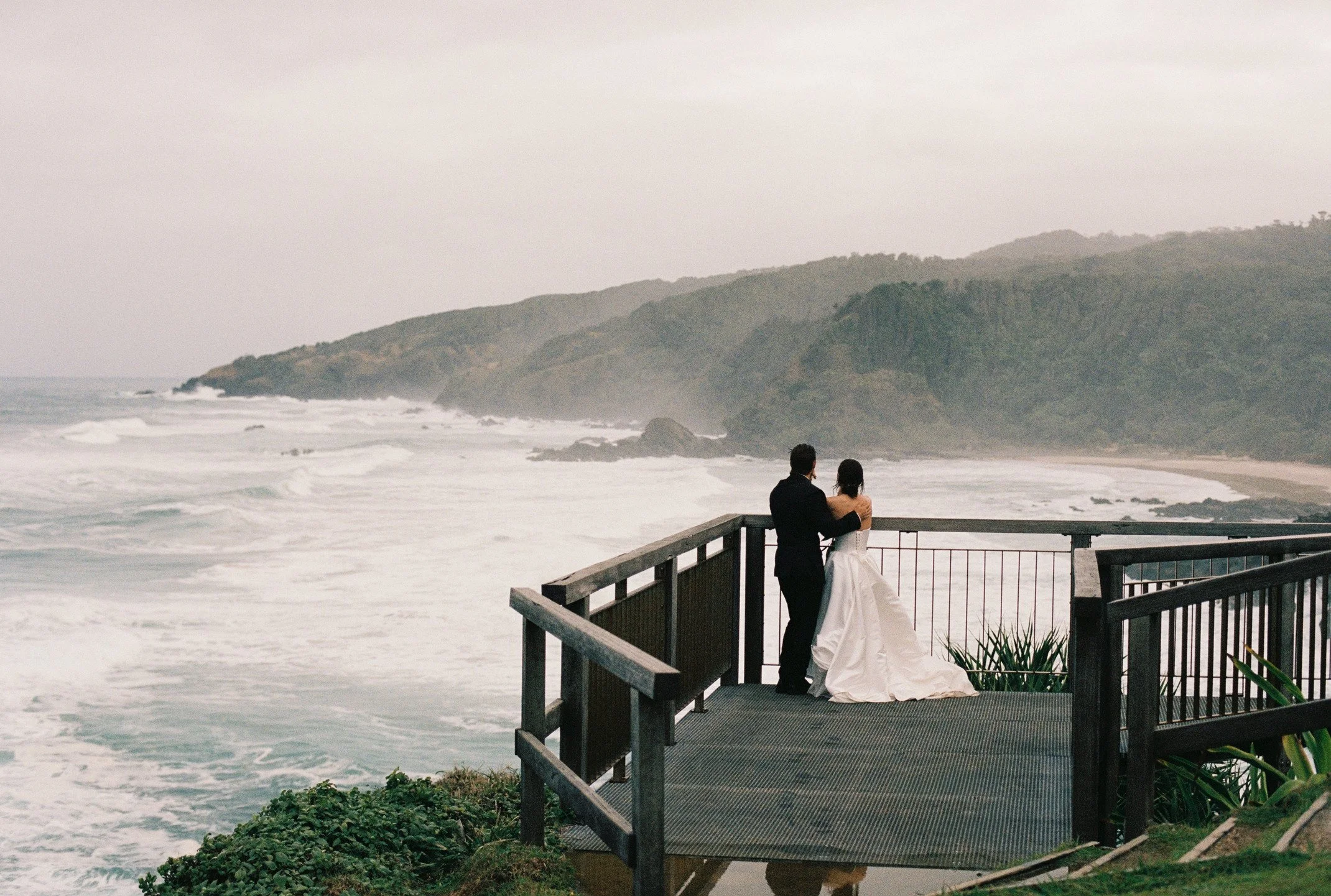 A couple in wedding attire standing on a balcony overlooking an ocean with mountains in the background.
