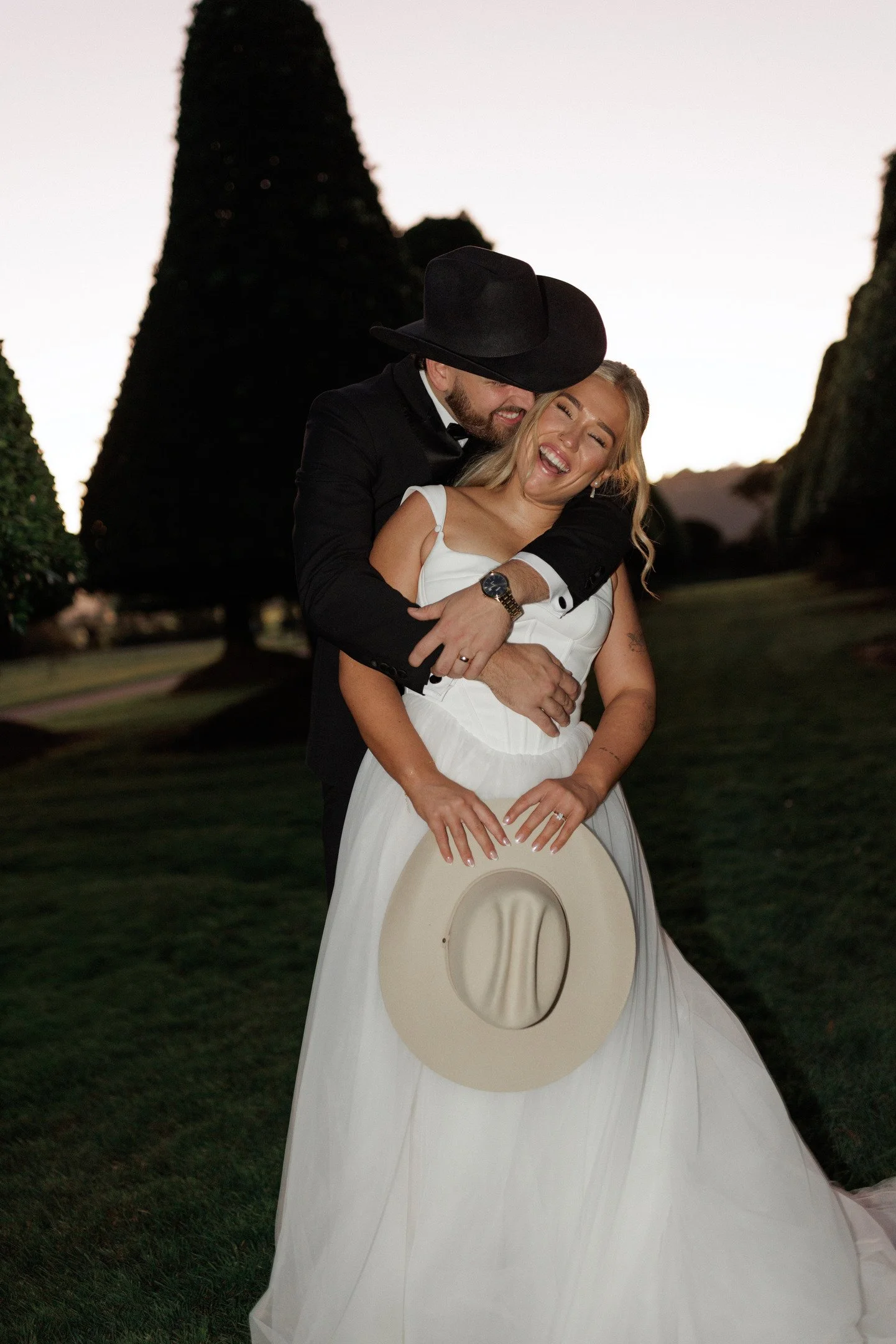 A couple dressed in wedding attire sharing a joyful embrace outdoors during dusk, with trees and a clear sky in the background.