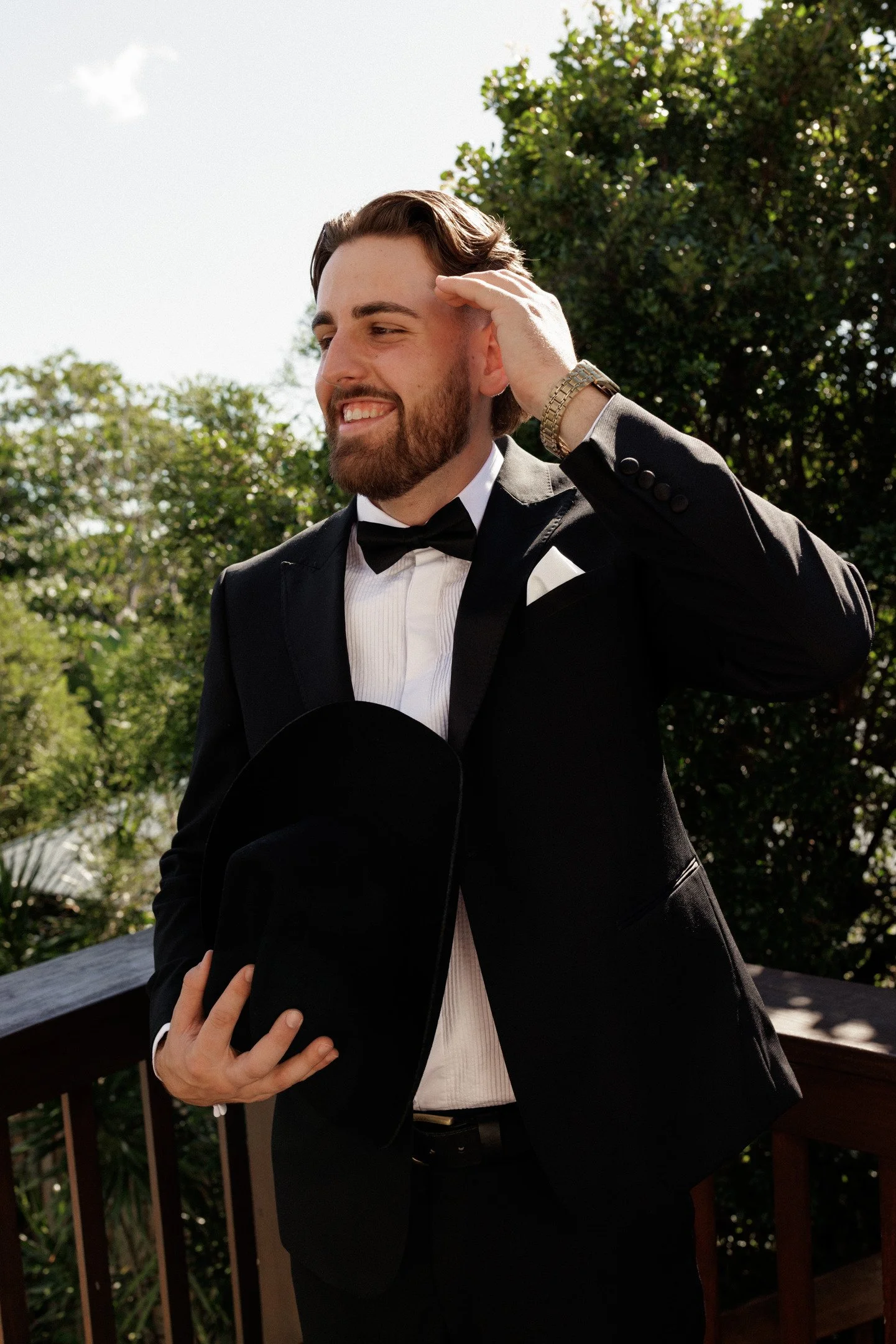 A man in a tuxedo with a bow tie, holding a black hat, smiling, and scratching his head outdoors with trees and a wooden railing in the background.