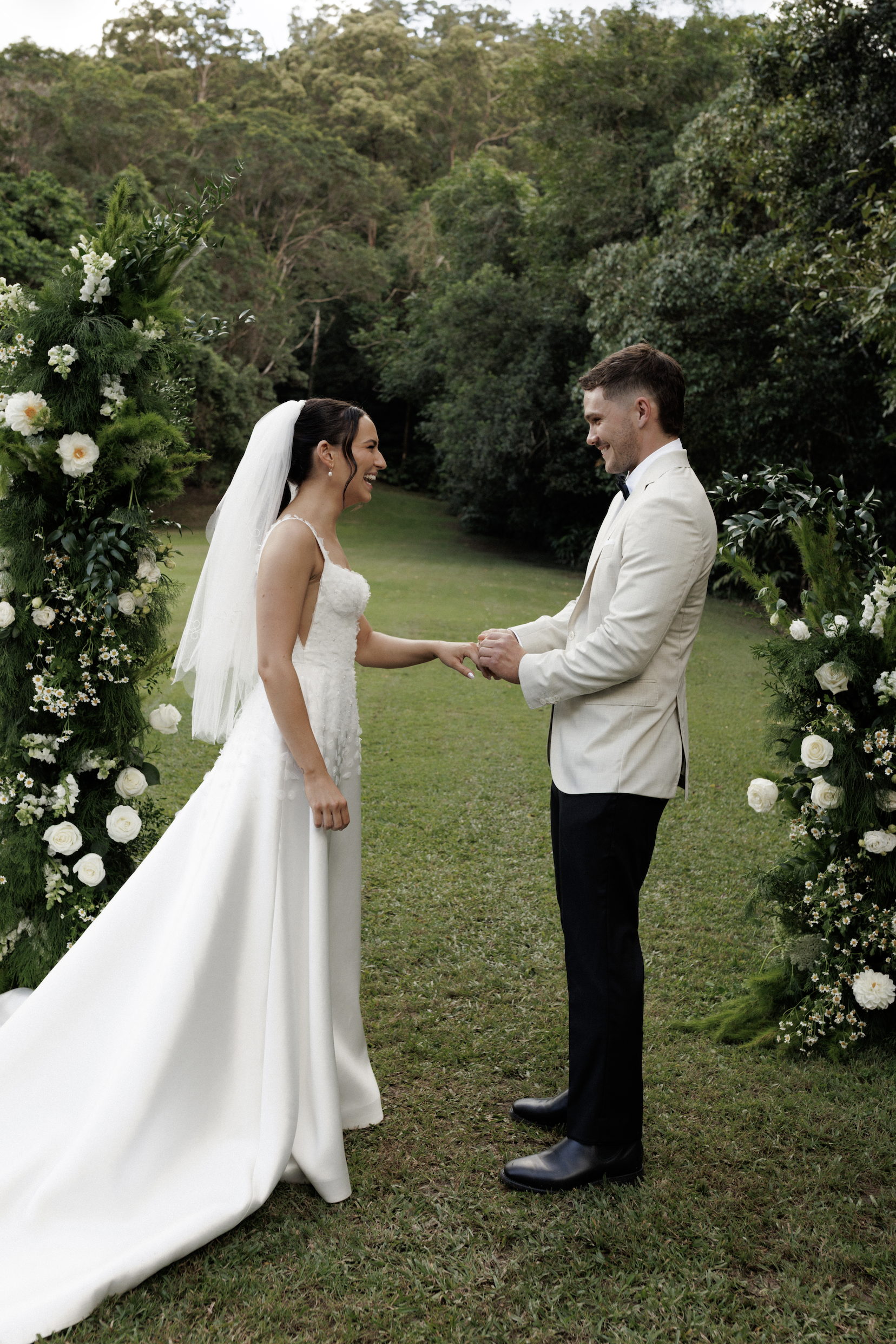 A bride and groom standing outdoors during a wedding ceremony, holding hands and smiling at each other, surrounded by greenery and floral arrangements.
